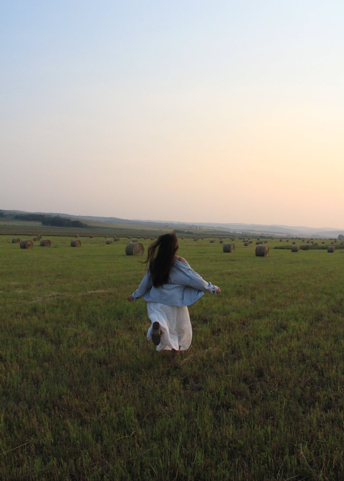 A woman in a light blue denim jacket and white skirt runs joyfully across a grassy field with hay bales under a soft sunset sky.