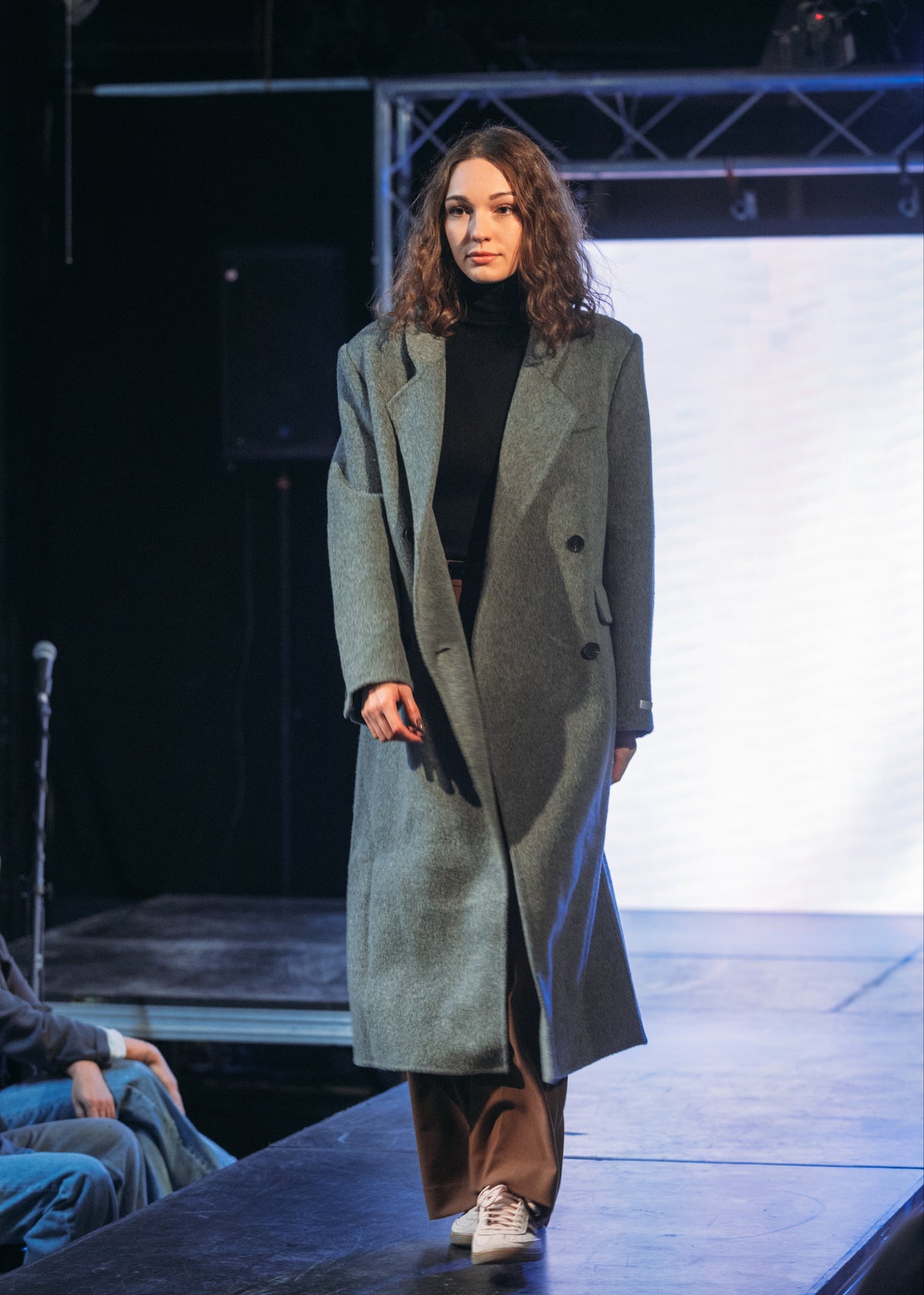 A model walks a fashion runway wearing a long, double-breasted grey coat over a black turtleneck and brown wide-leg pants.