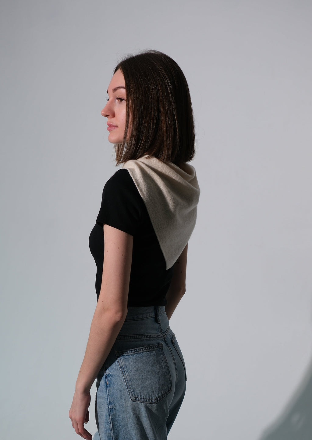 A woman in profile wearing a black short-sleeve top with a beige draped shawl, paired with wide-leg blue jeans, standing against a light gray studio background with a prominent shadow.