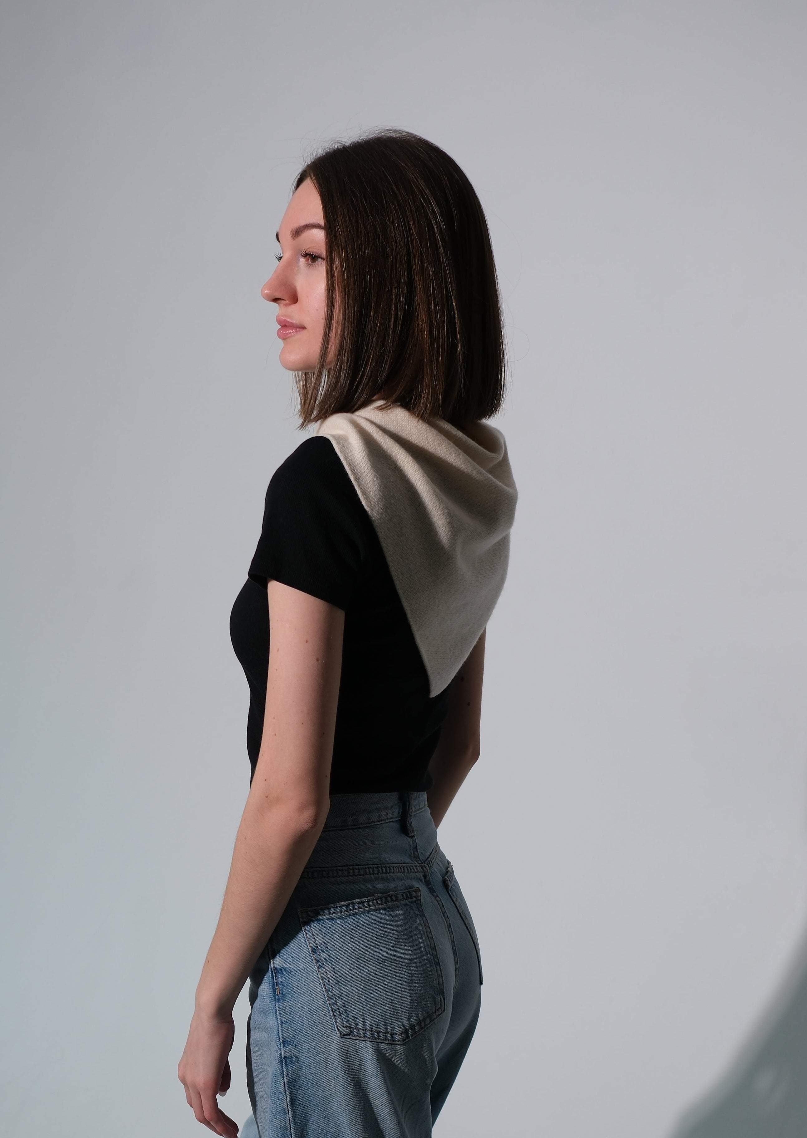 A woman in profile wearing a black short-sleeve top with a beige draped shawl, paired with wide-leg blue jeans, standing against a light gray studio background with a prominent shadow.