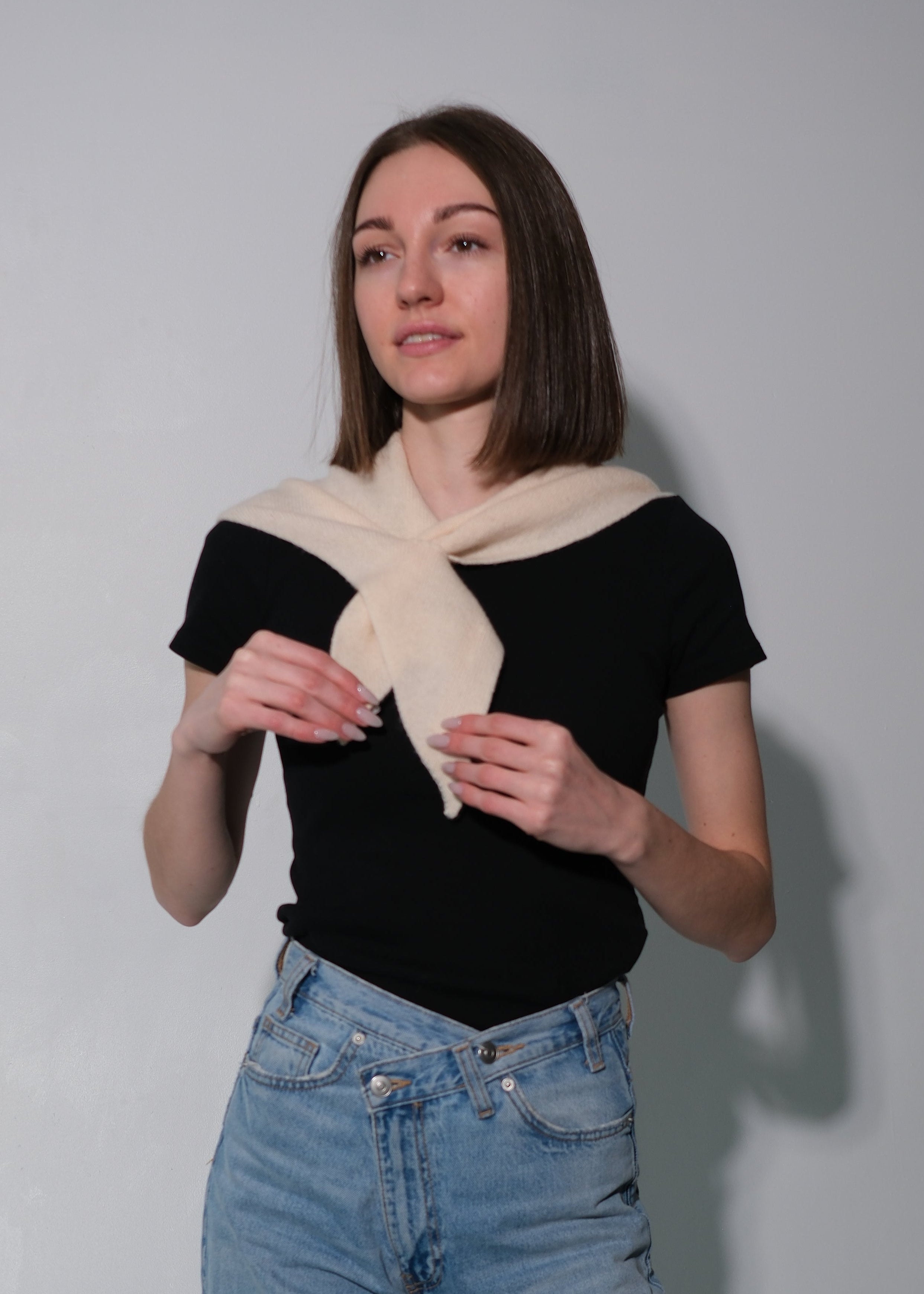 A woman modeling a black short-sleeve top with a cream-colored knit scarf tied around her neck, paired with light-wash blue jeans, standing against a plain gray wall.