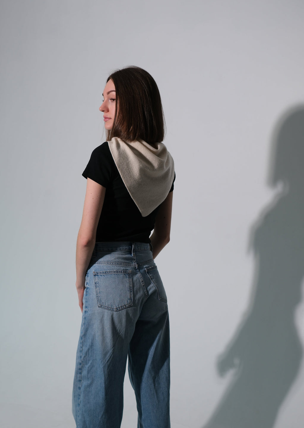 A woman stands in profile wearing a black short-sleeve top, a beige shawl draped over her shoulders, and wide-leg blue jeans against a plain light gray studio wall, with a long shadow cast to the right.