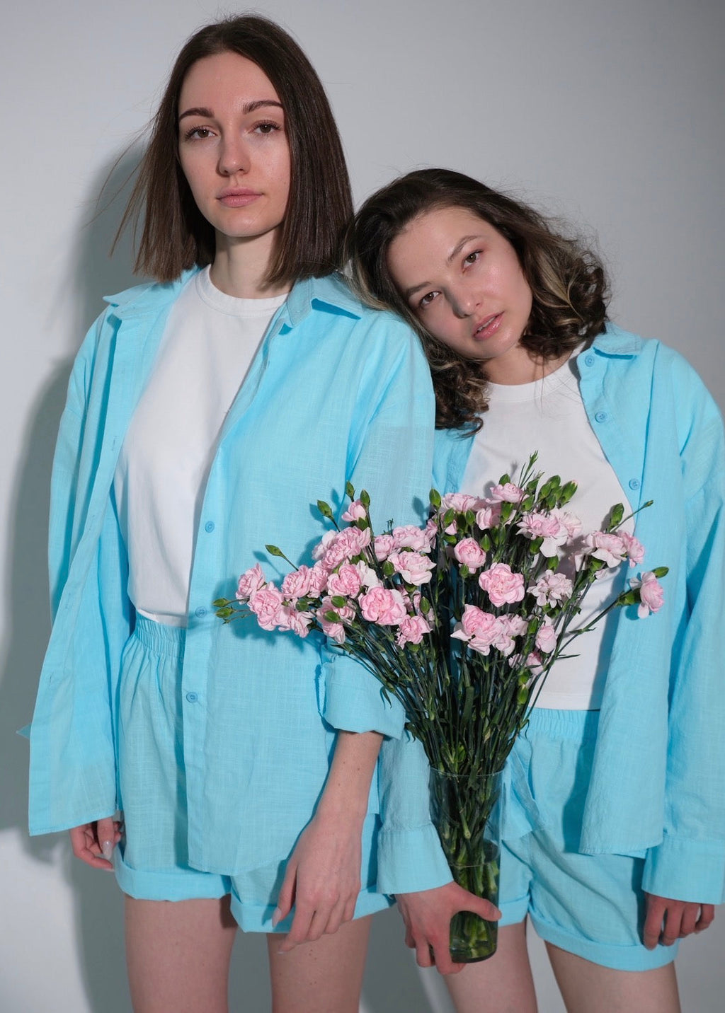 Two people wearing matching light blue outfits holding pink flowers against a plain background