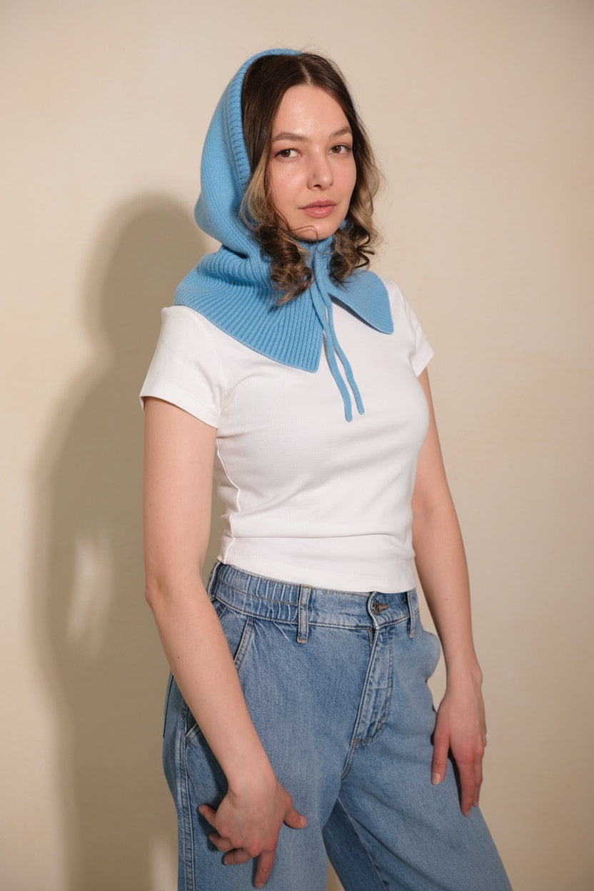 Person wearing a blue knitted hood over a white shirt and blue jeans against a gray background