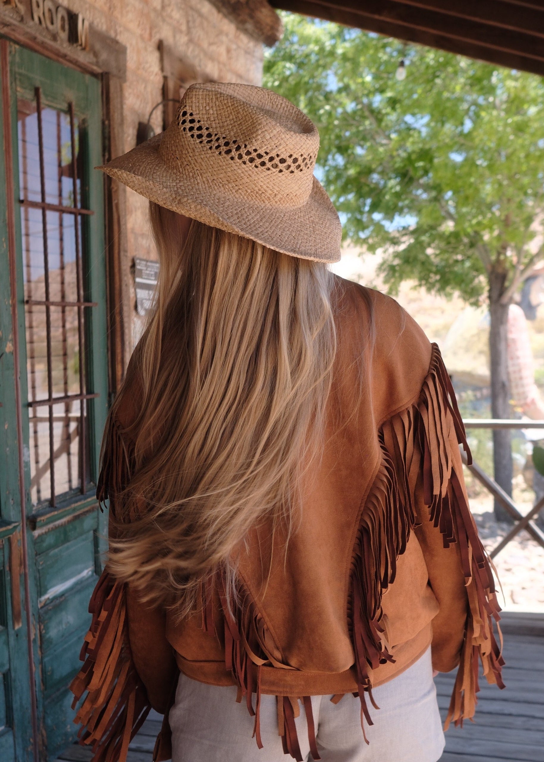 Person wearing a brown fringed jacket and hat in front of a rustic building.