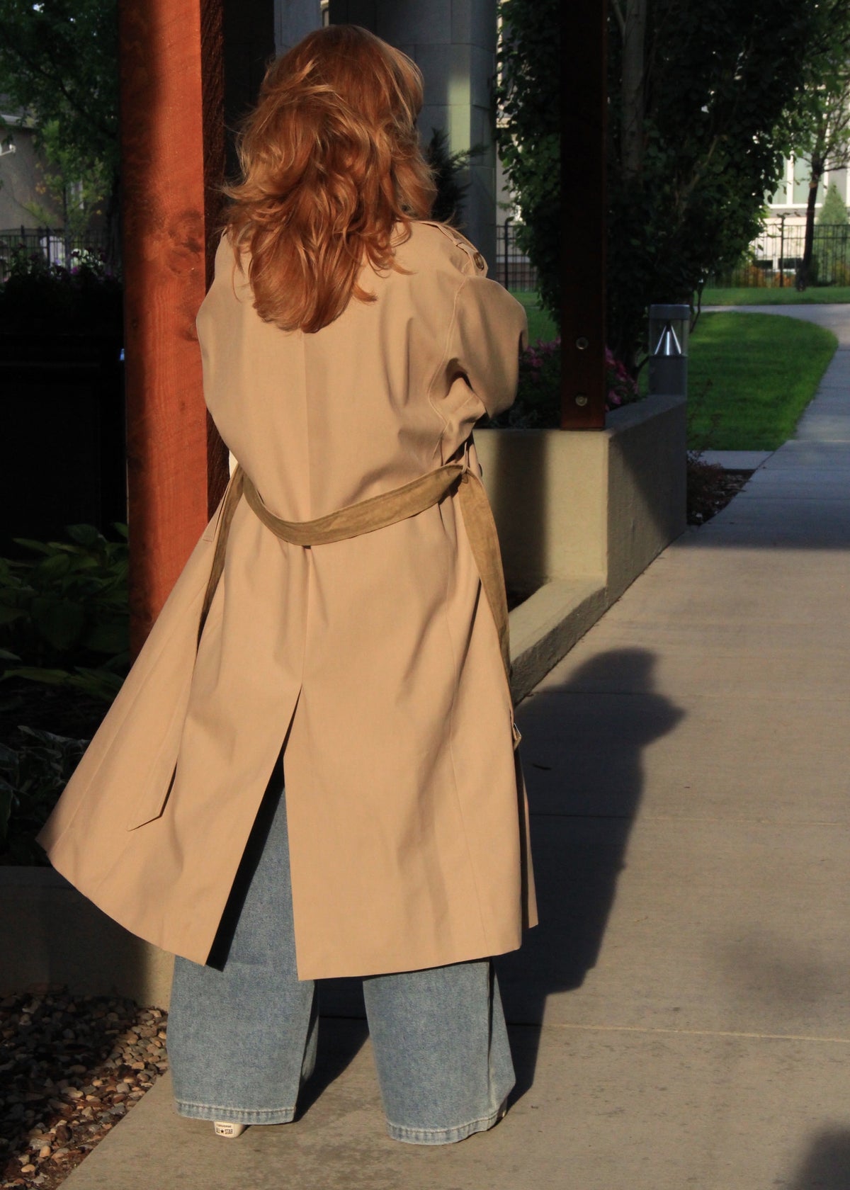 A woman viewed from behind wearing a beige trench coat with a matching belt, paired with wide-leg blue jeans, standing on a concrete walkway outdoors.