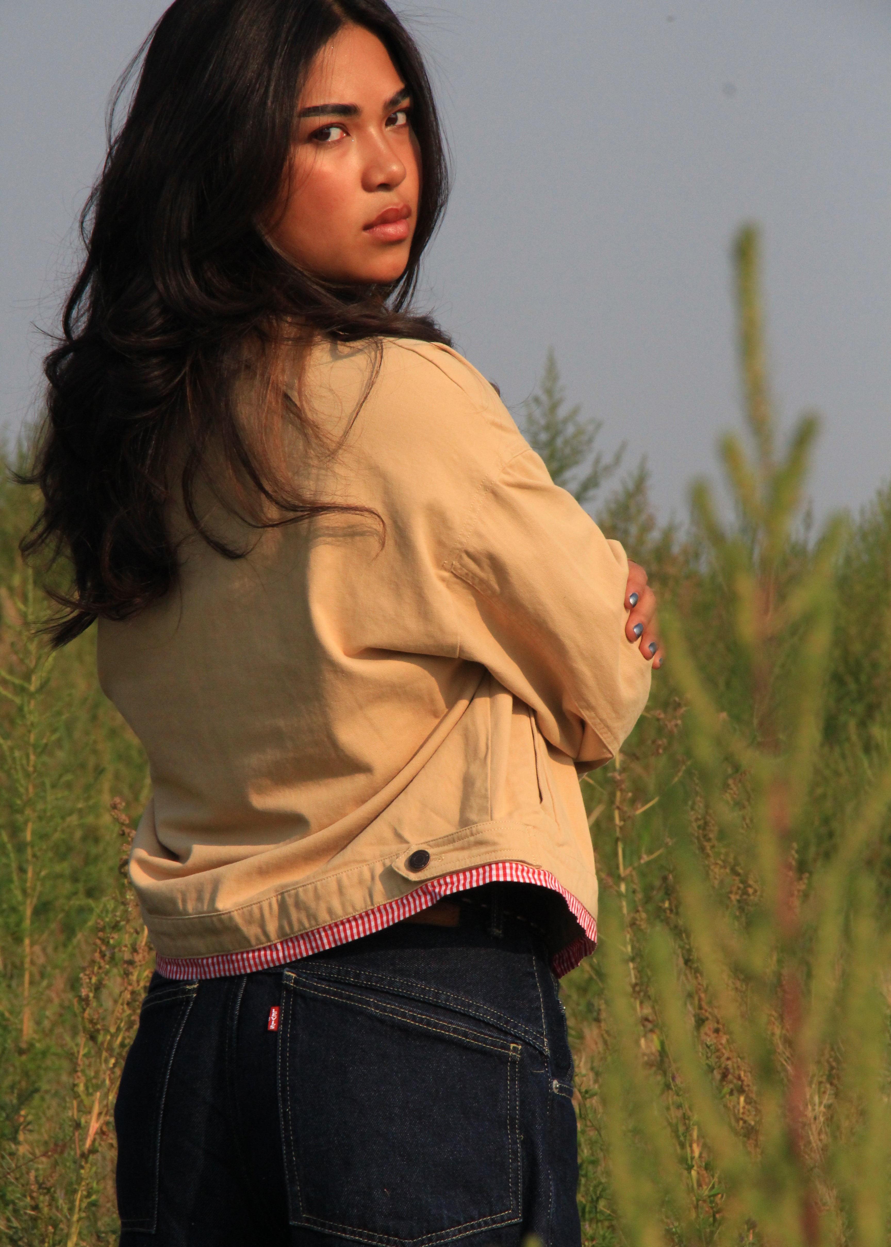 A woman wearing a beige utility jacket over a pink striped shirt and dark blue jeans, standing in a grassy outdoor field.