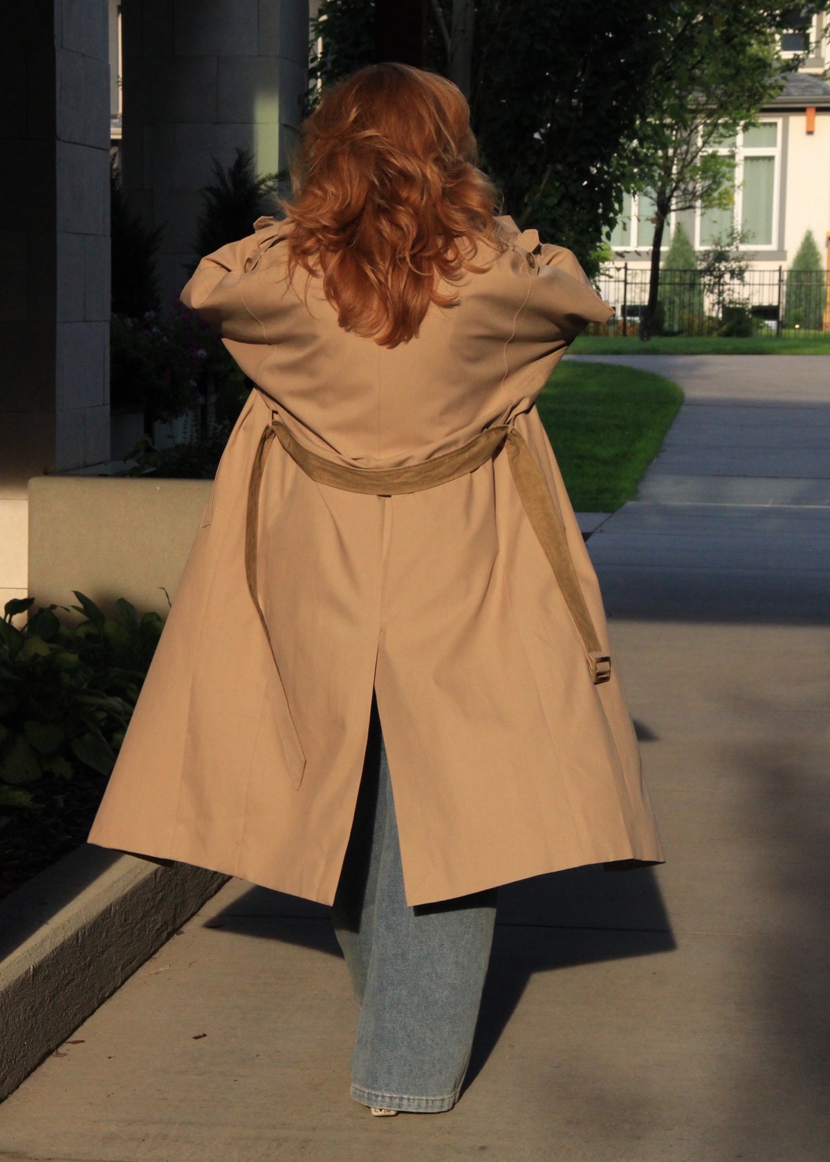 A woman with long red hair is shown from behind wearing a beige trench coat over light blue jeans, standing on a concrete sidewalk outdoors.