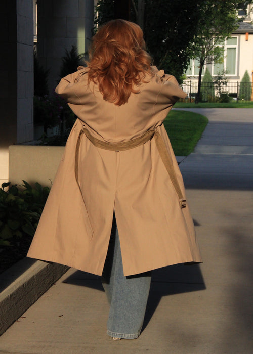 A woman with long red hair is shown from behind wearing a beige trench coat over light blue jeans, standing on a concrete sidewalk outdoors.