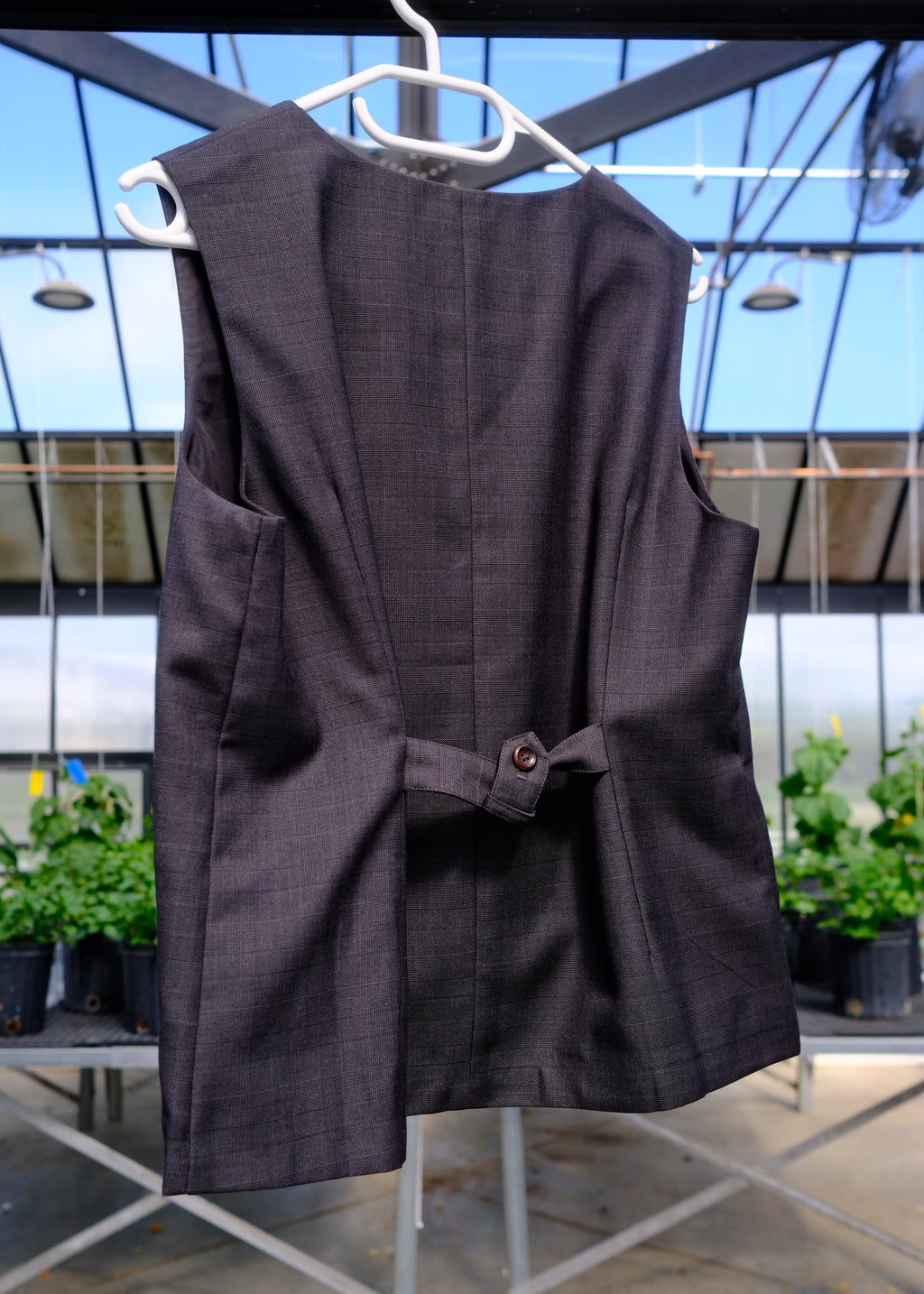 A dark brown sleeveless vest with a small button detail at the waist, hanging on a white plastic hanger against a glass window background.
