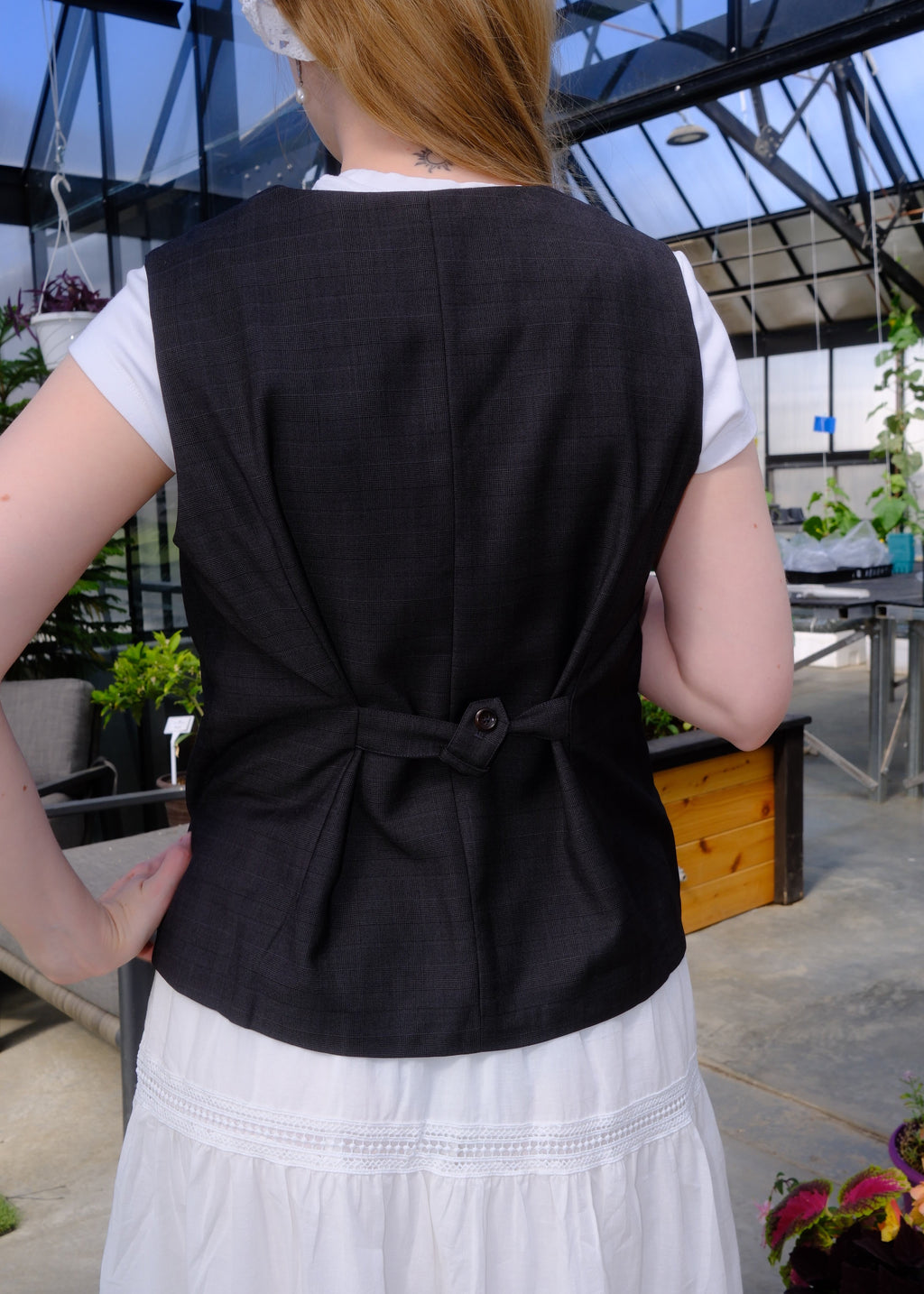 A person wearing a black structured vest over a white top, photographed from the back in an outdoor greenhouse or garden setting with plants and glass panels visible.