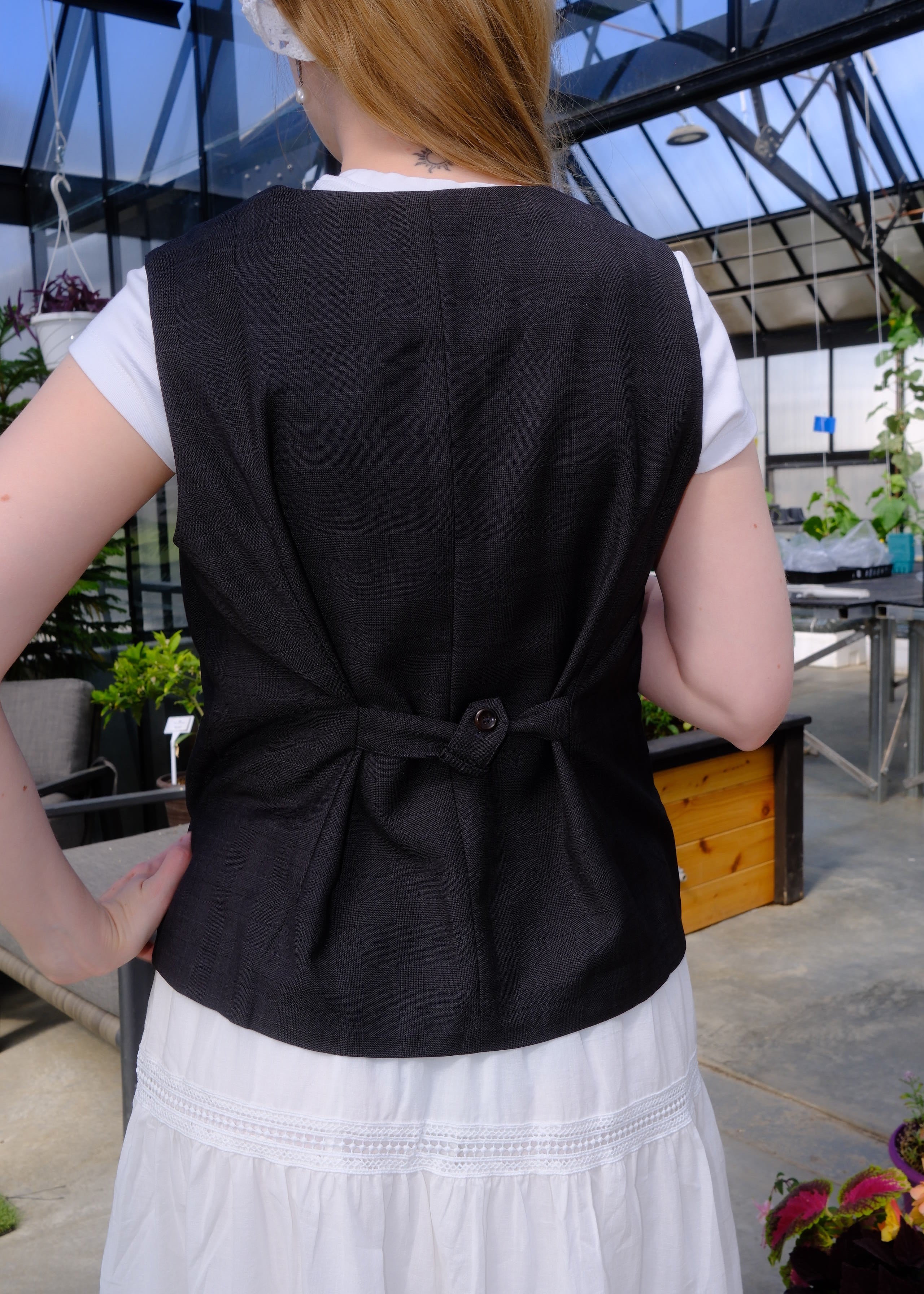 A person wearing a black structured vest over a white top, photographed from the back in an outdoor greenhouse or garden setting with plants and glass panels visible.