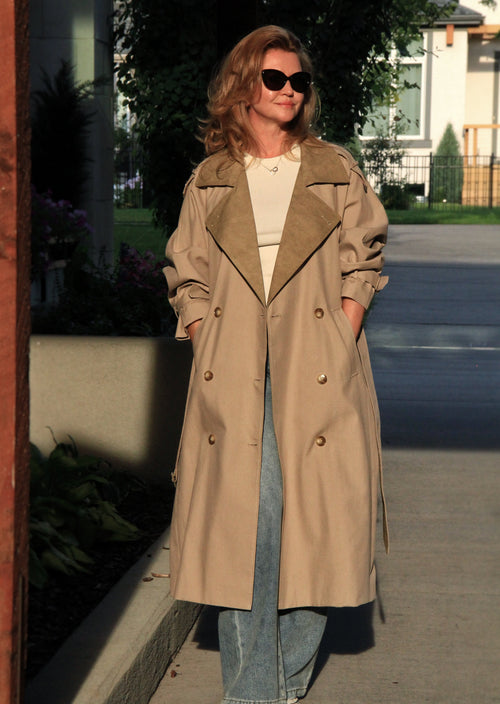 A woman wearing a long beige trench coat over a cream top and blue jeans, standing outdoors on a sidewalk in front of a house.
