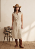 Woman wearing a beige sleeveless dress, brown cowboy hat, and boots on a wooden floor. Western style outfit in Calgary