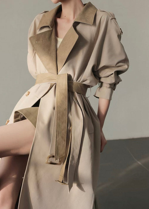 A model wearing a beige trench coat with a wide collar, belted waist, and voluminous sleeves, photographed in a minimalist studio setting.