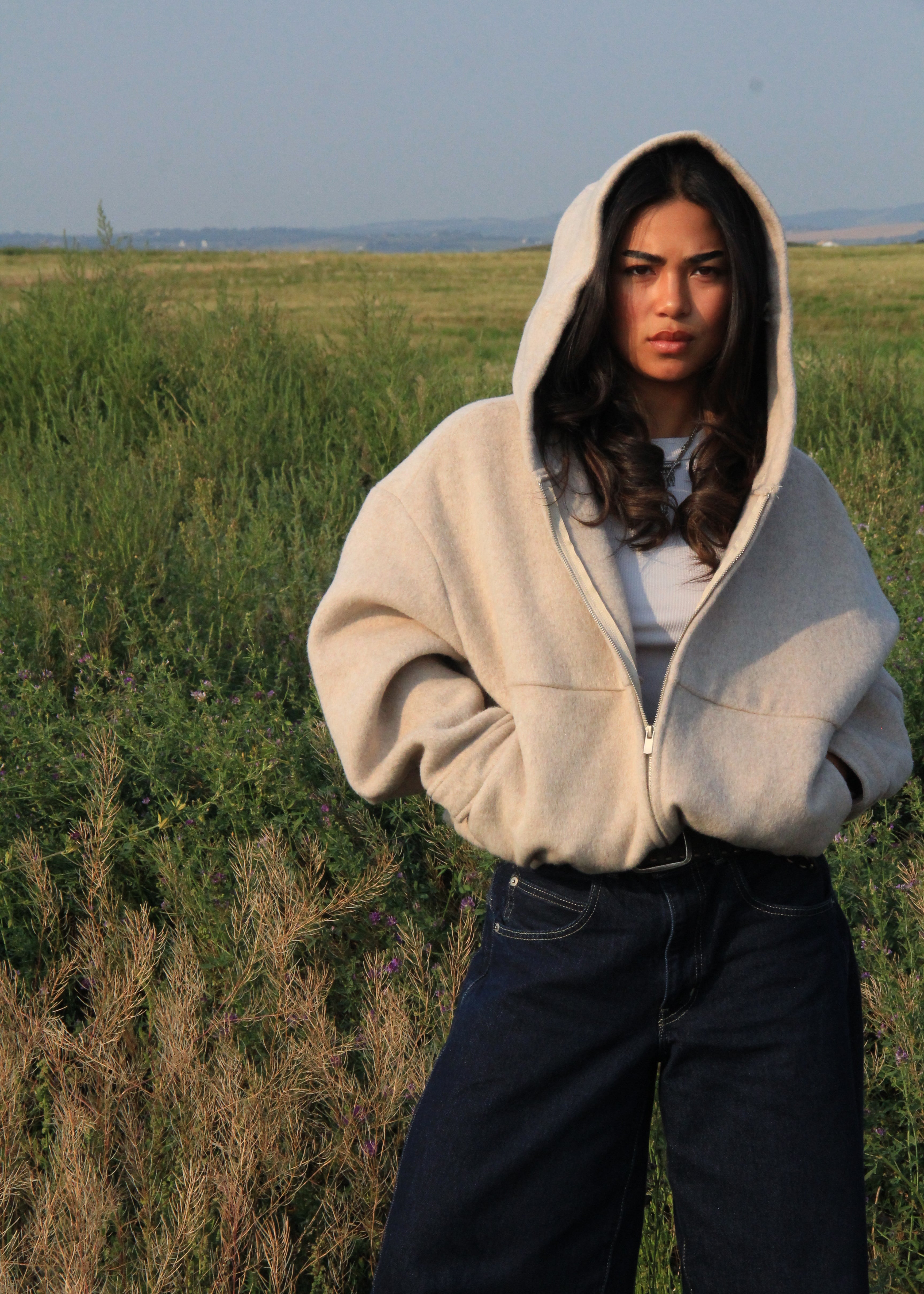 A woman wearing a cream-colored oversized zip-up hoodie and dark wide-leg jeans, standing in a grassy field.