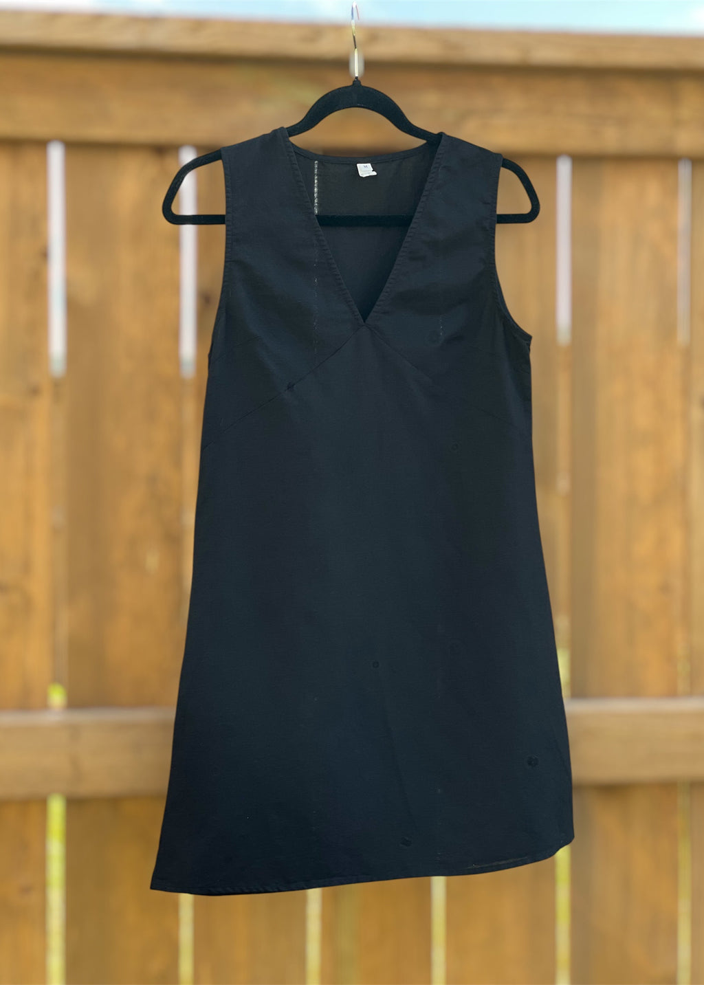 A black sleeveless V-neck dress hanging on a black hanger against a wooden fence background.