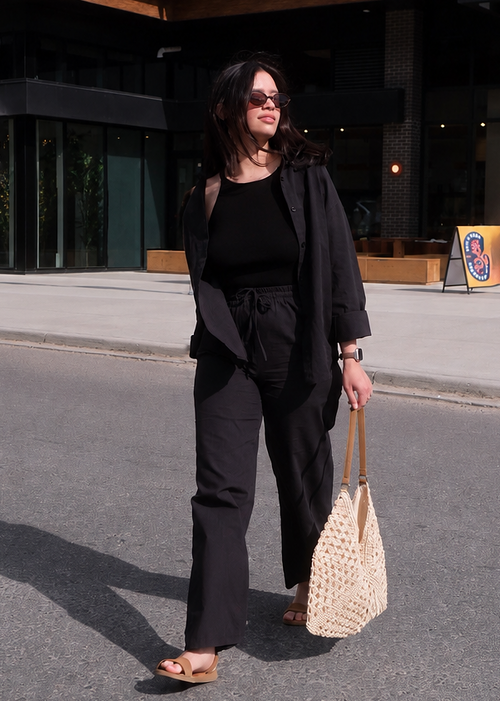 Woman in black outfit holding a woven bag on a city street.