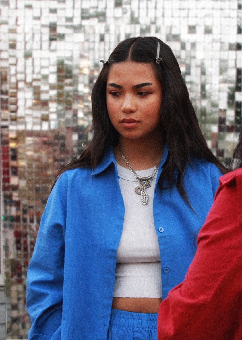 A woman wearing a bright blue button-up shirt over a white crop top, standing in front of a reflective mirrored wall. A red garment is partially visible on the right edge.