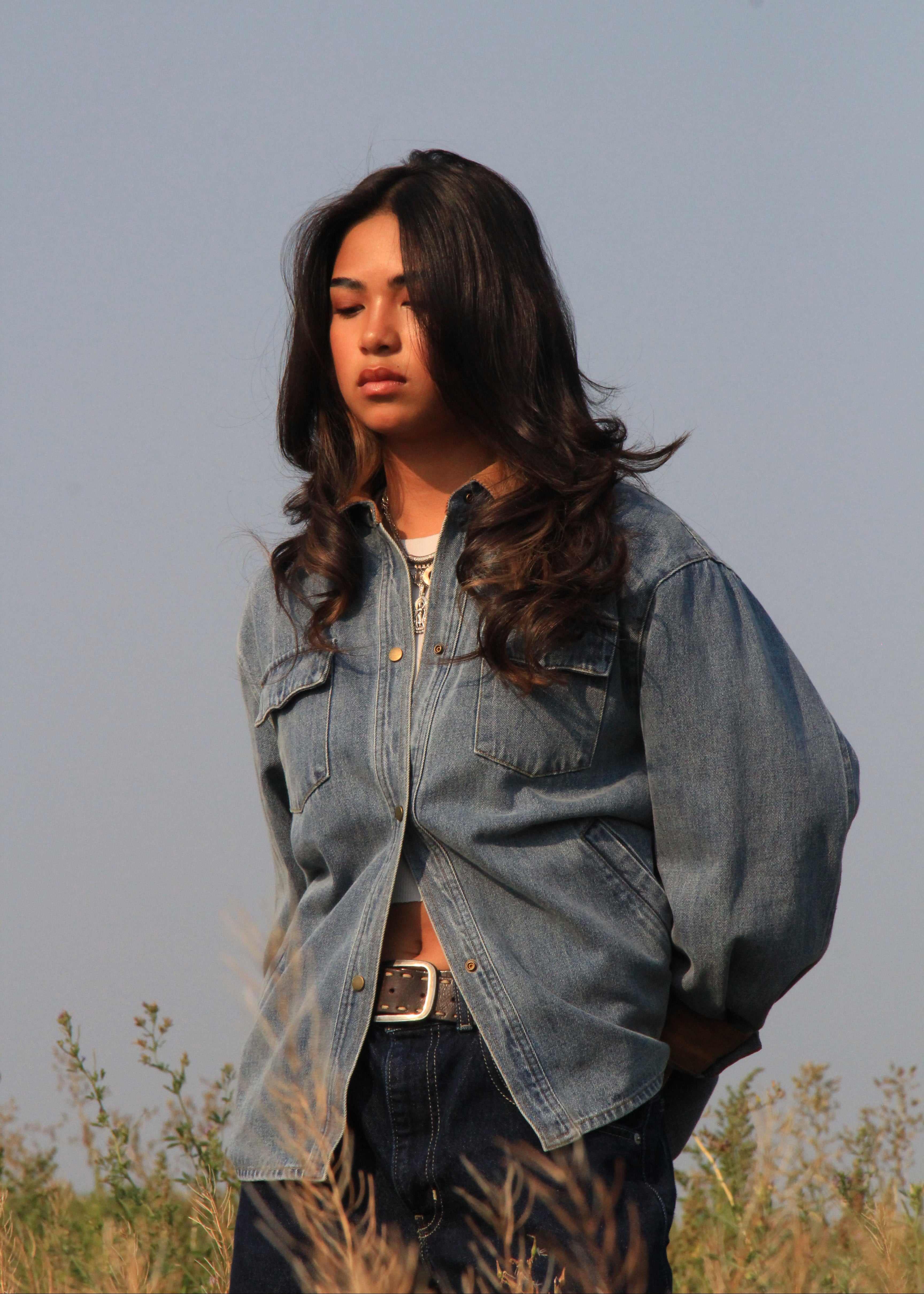 A woman standing outdoors in a field, wearing an oversized light blue denim button-up shirt over a white top and dark jeans.