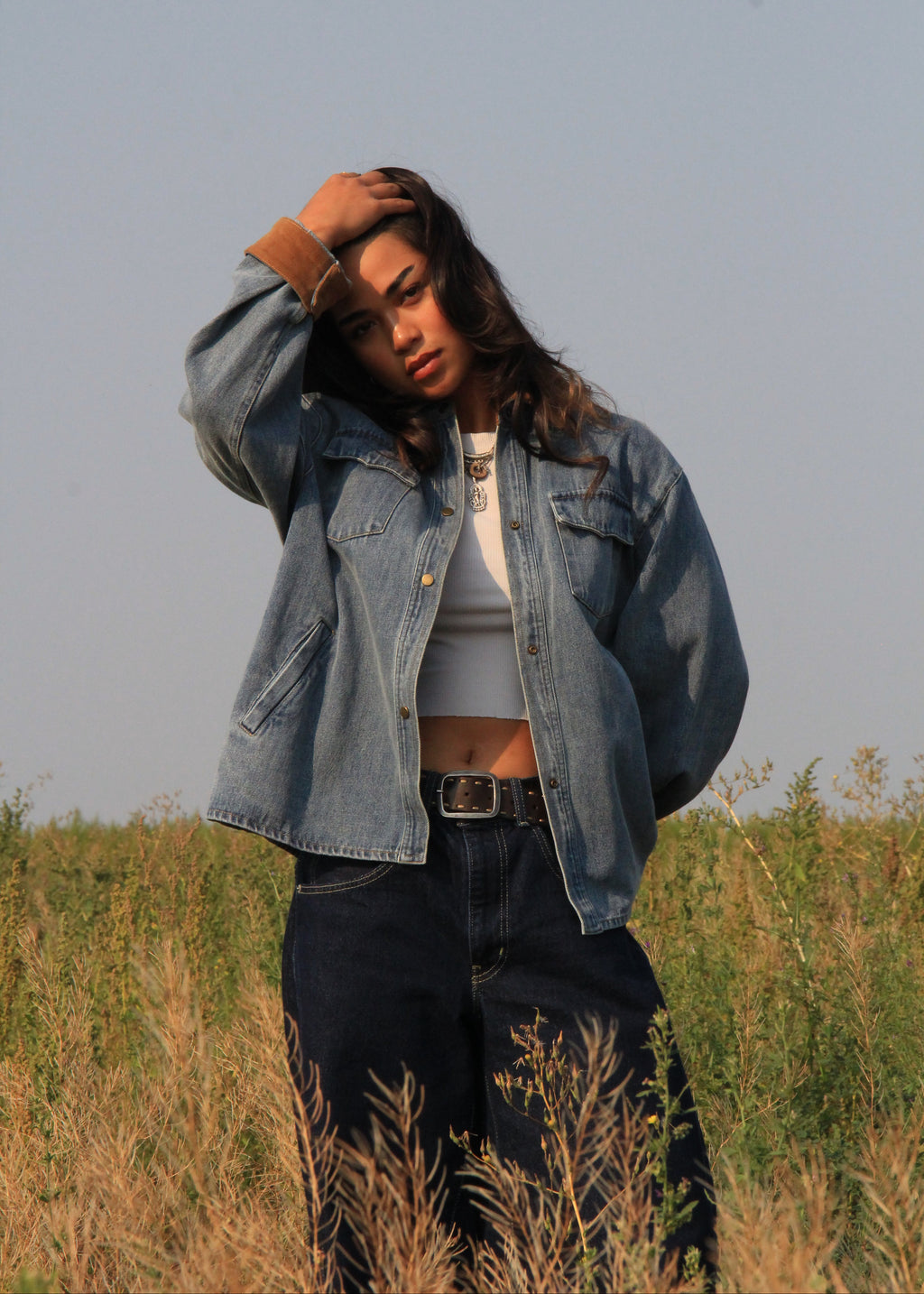 A woman standing in a grassy field wearing an oversized light blue denim jacket over a white crop top and dark jeans.