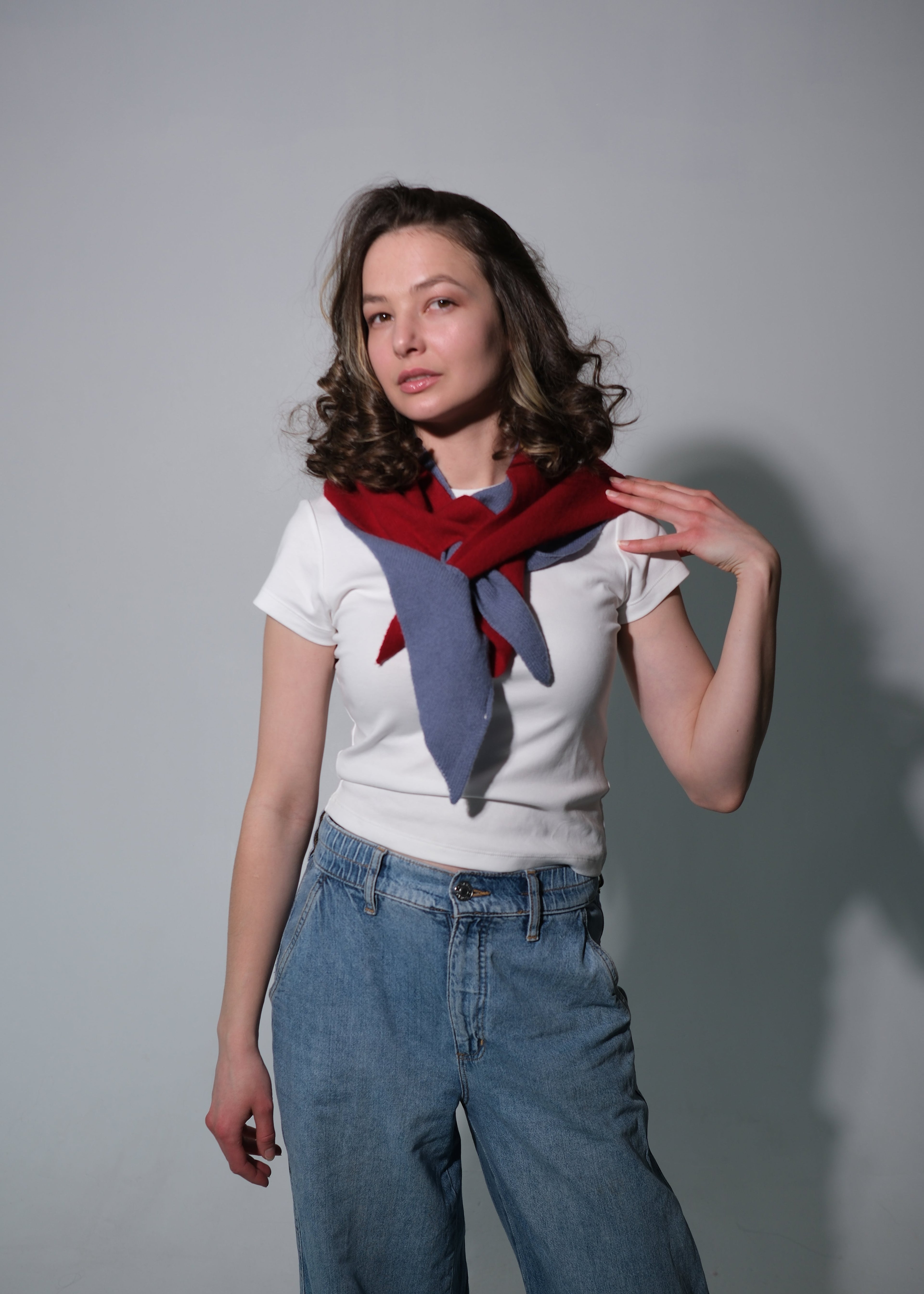 A woman modeling a white t-shirt and blue jeans, wearing a red and blue knit scarf tied around her neck, against a plain gray studio background.