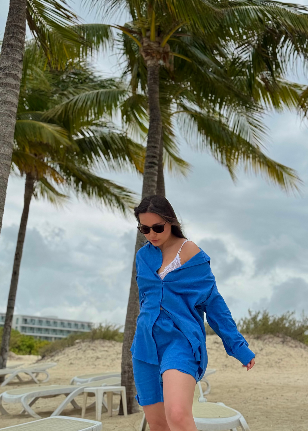 A woman wearing a bright blue matching lounge set walks on a sandy beach with palm trees and lounge chairs in the background.