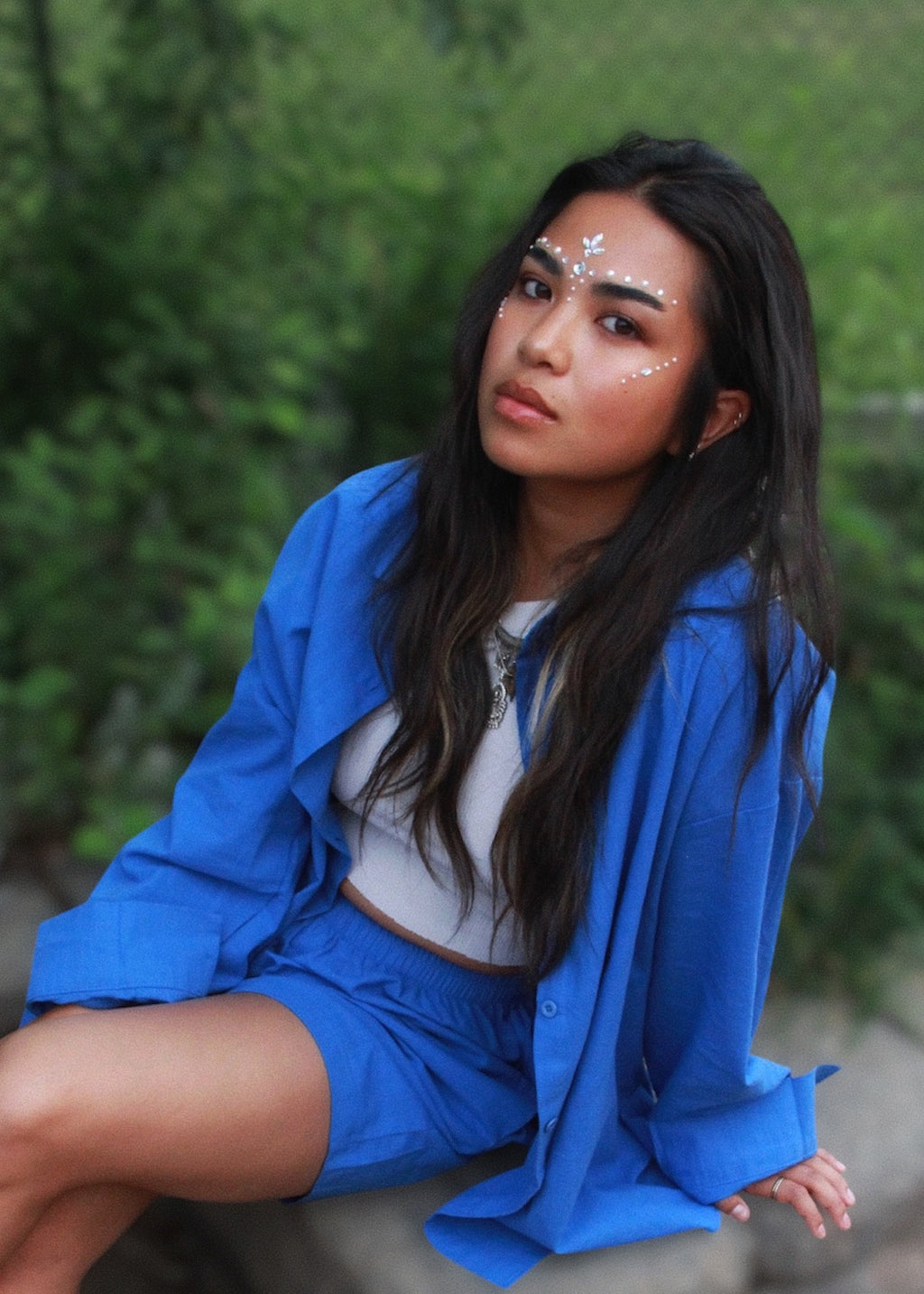 A woman with white glitter face makeup sits outdoors wearing a bright blue button-up shirt and matching blue shorts over a white crop top, with a blurred green foliage background.
