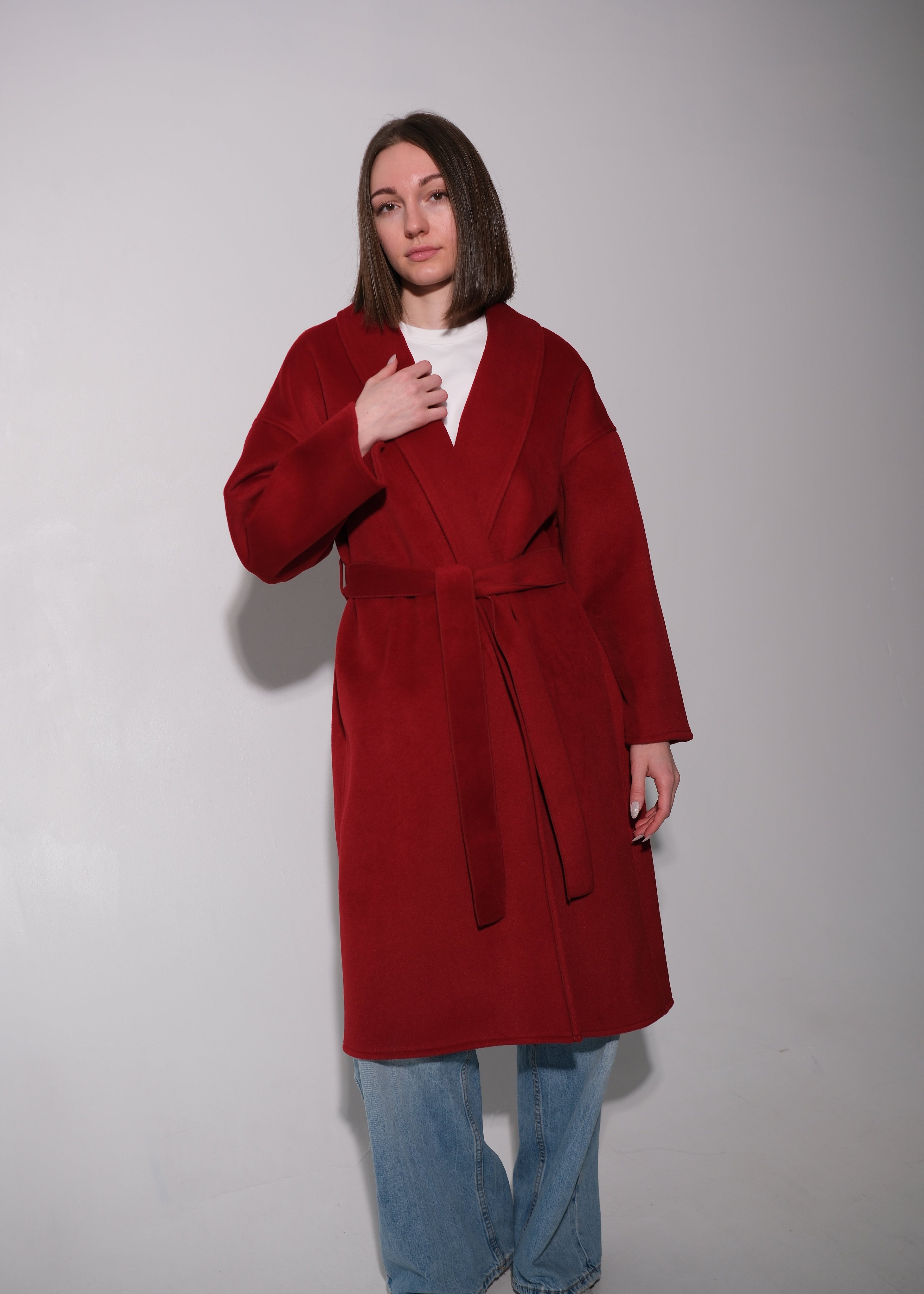 A woman modeling a long, deep red wool-blend coat with a matching tie belt, worn over a white top and blue jeans, standing against a plain light gray studio background.