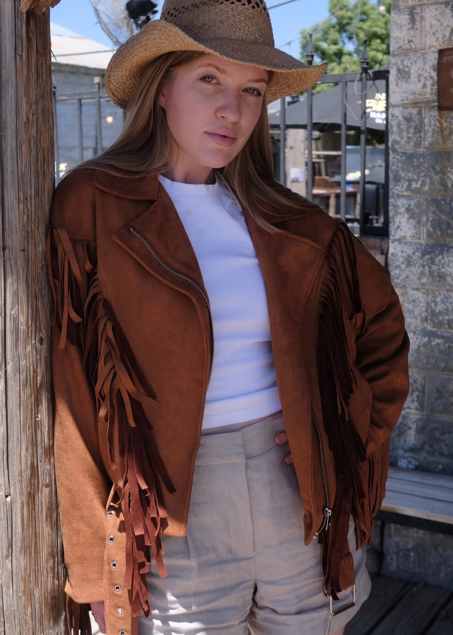 Woman wearing a western-inspired brown fringe jacket over a white fitted top and beige shorts, photographed on a rustic wooden porch. The suede-look jacket features long fringe details and a biker-style silhouette, creating a classic Calgary Stampede and cowboy fashion aesthetic.