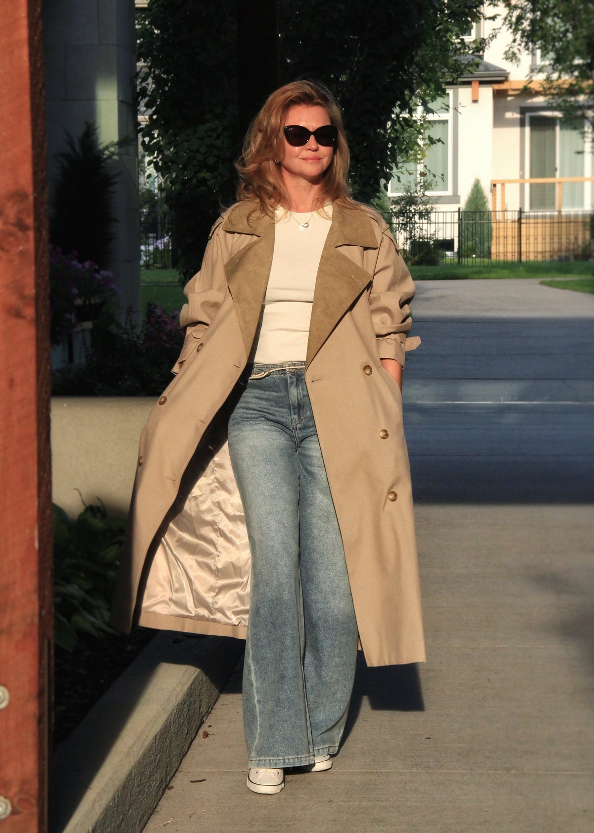 A woman walking outdoors on a sunny day wearing a long beige trench coat over a white top and light blue flared jeans.