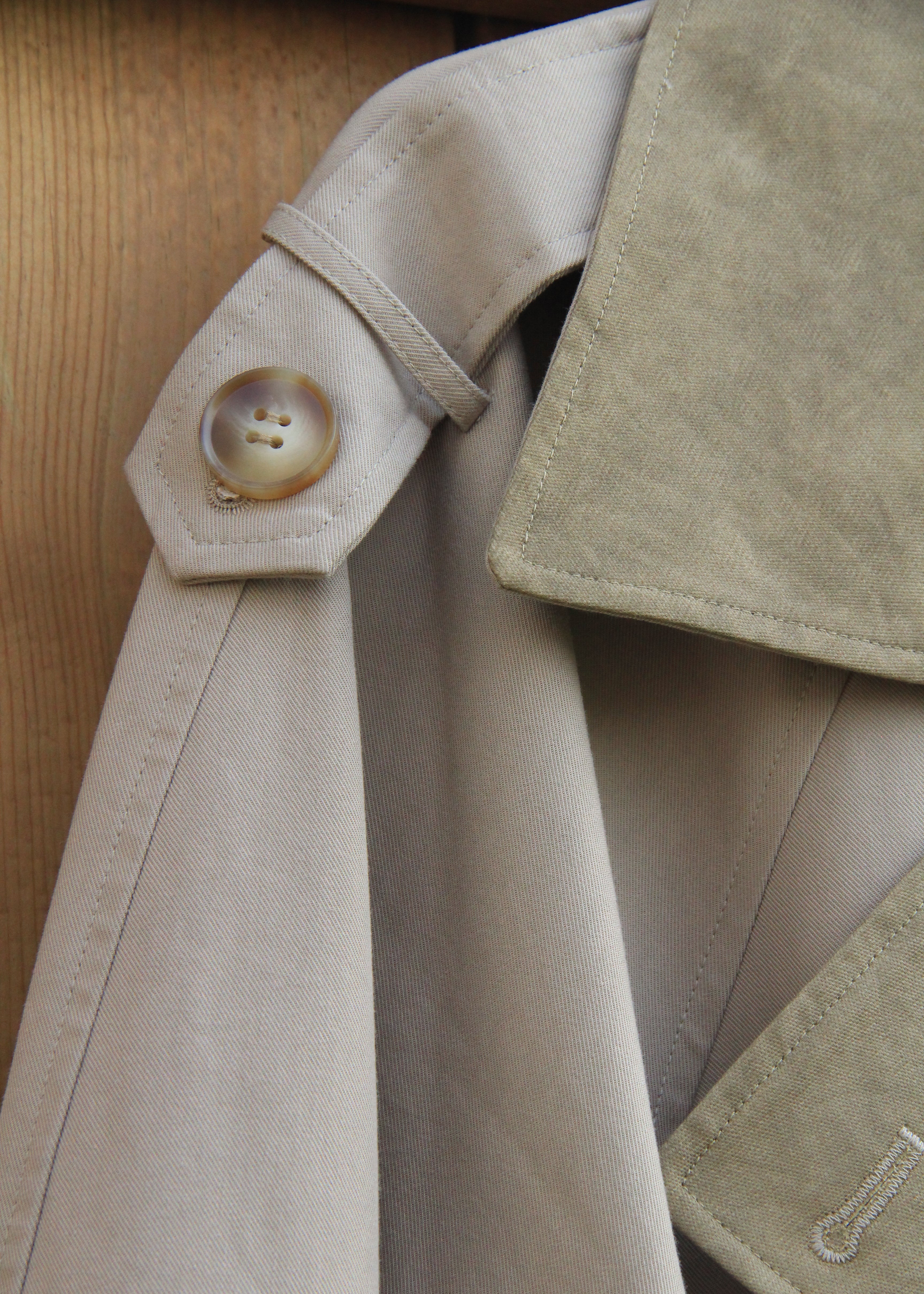 Close-up of a beige trench coat showing the shoulder tab with a round button, collar, and fabric texture against a wooden background.