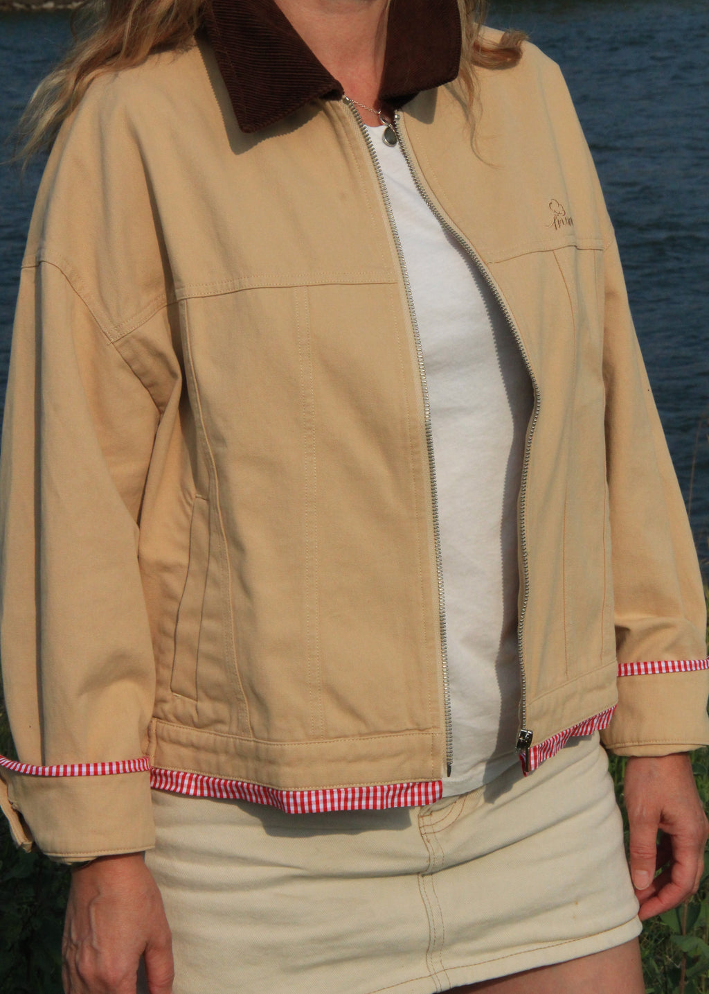 A woman wearing a beige utility-style jacket with a dark brown corduroy collar and red-and-white gingham trim, layered over a white top and cream skirt, standing outdoors near water.