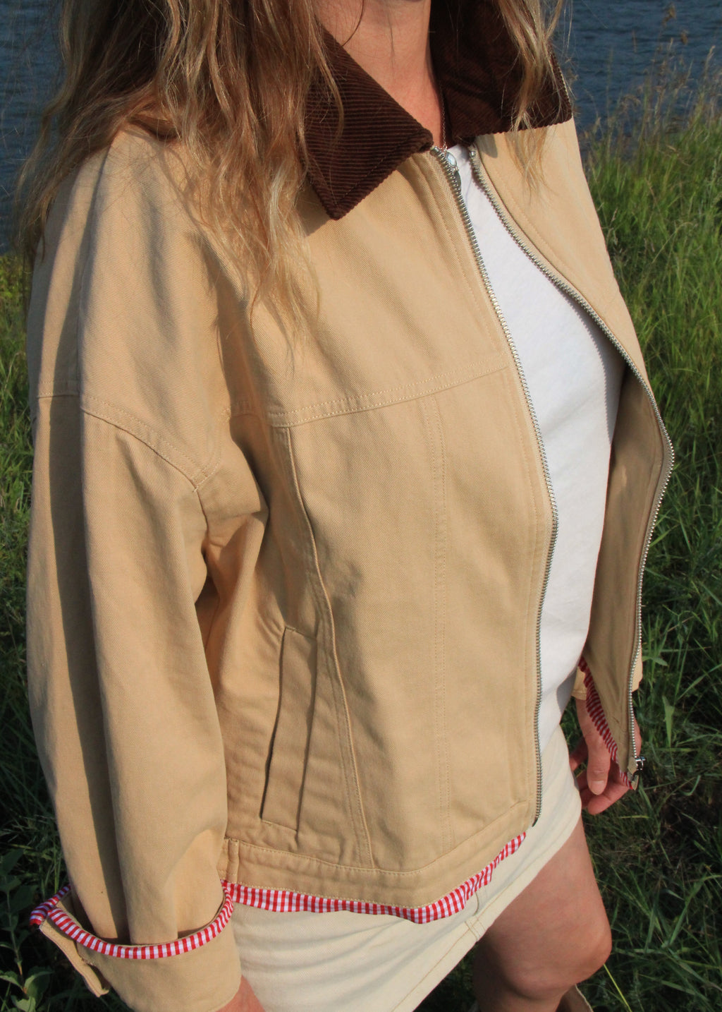 A woman wearing a beige utility-style jacket with a brown corduroy collar, zipped front, and red-and-white striped trim at the cuffs and hem, photographed outdoors near grass and water.