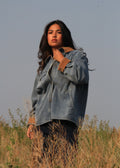 A woman wearing a light blue denim shirt jacket with brown suede-like cuffs stands in a field of dry grass under a clear sky.