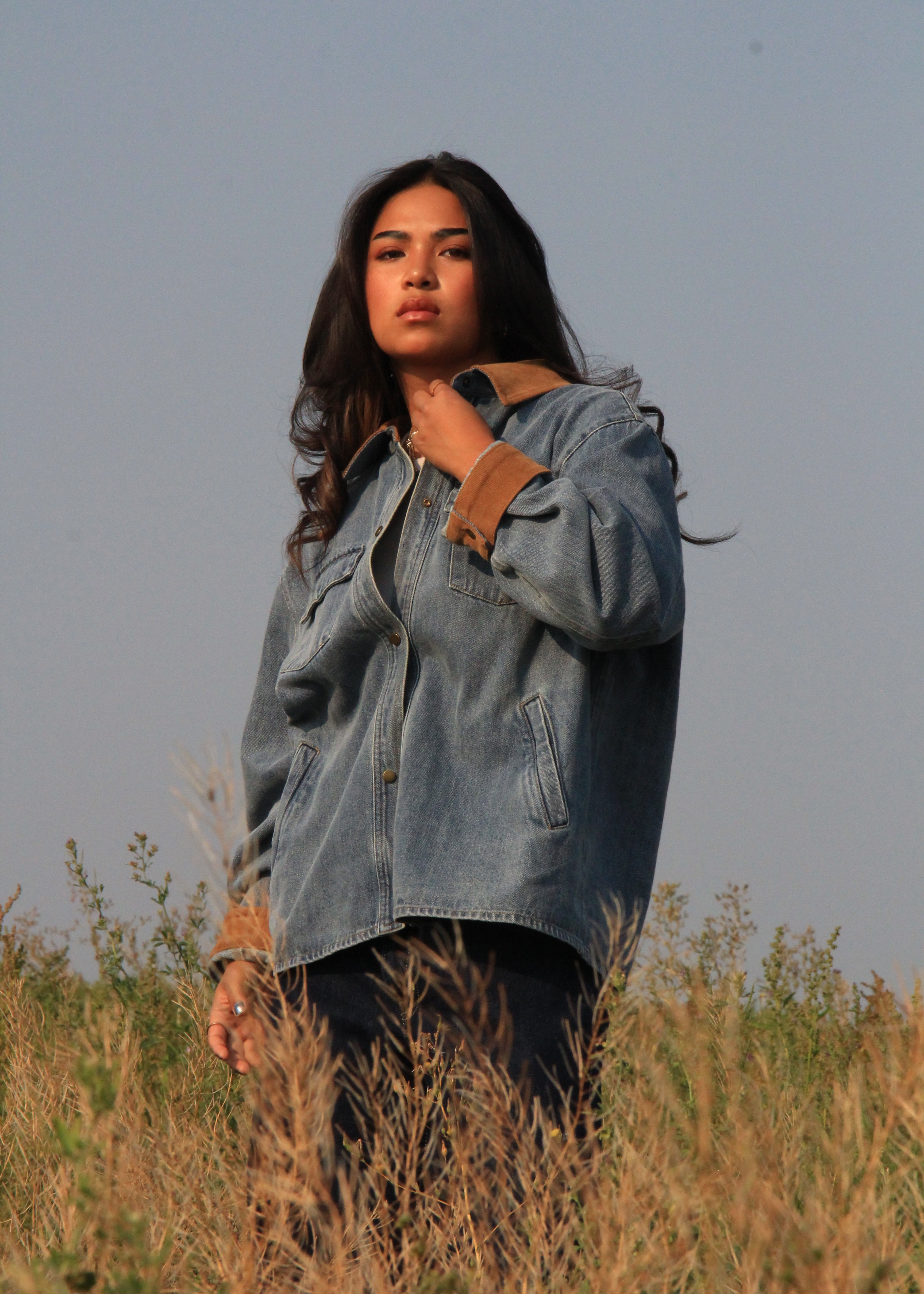 A woman wearing a light blue denim shirt jacket with brown suede-like cuffs stands in a field of dry grass under a clear sky.