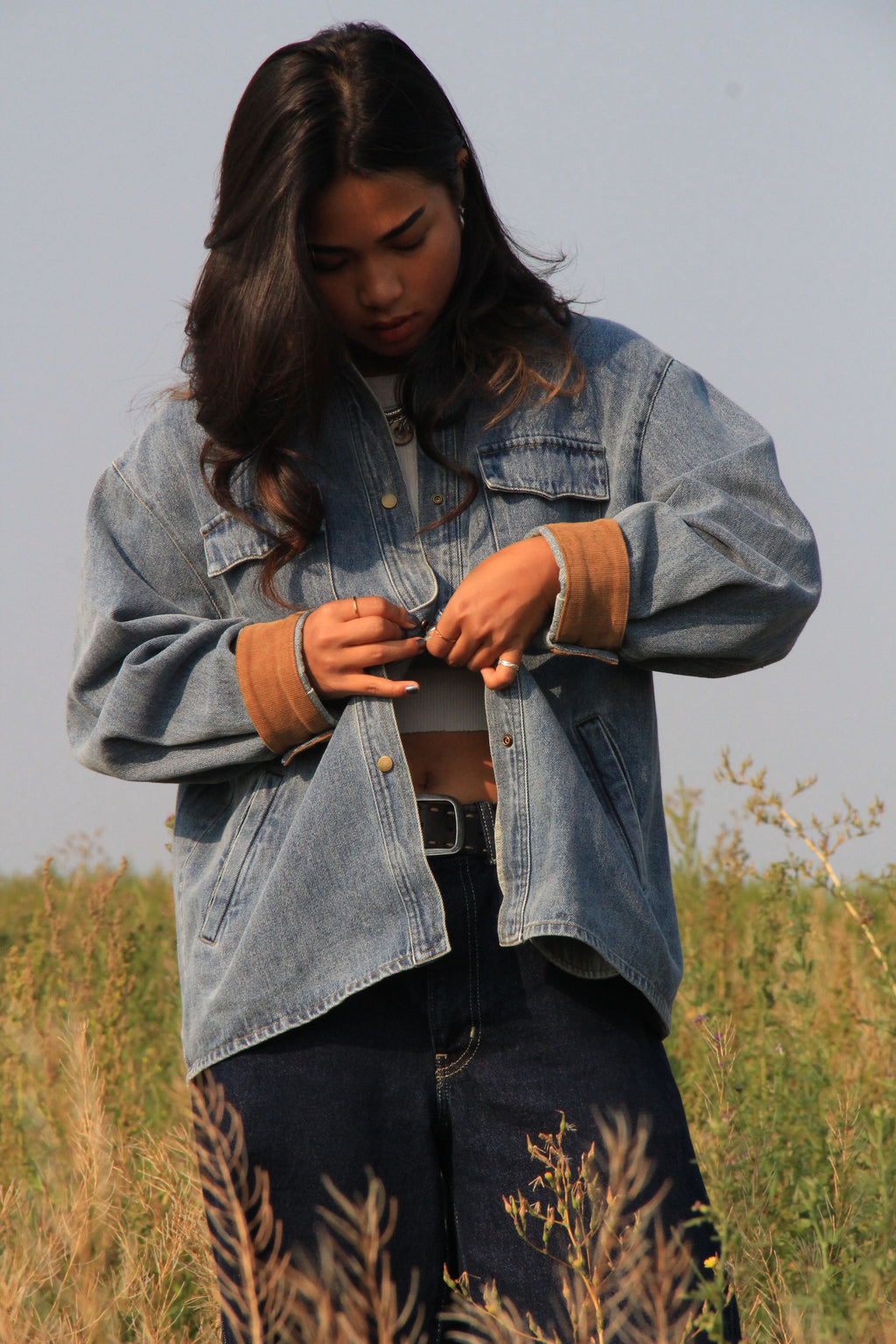 A woman standing in an outdoor grassy field wearing a light-wash blue denim jacket with tan ribbed cuffs, dark jeans, and a black belt.