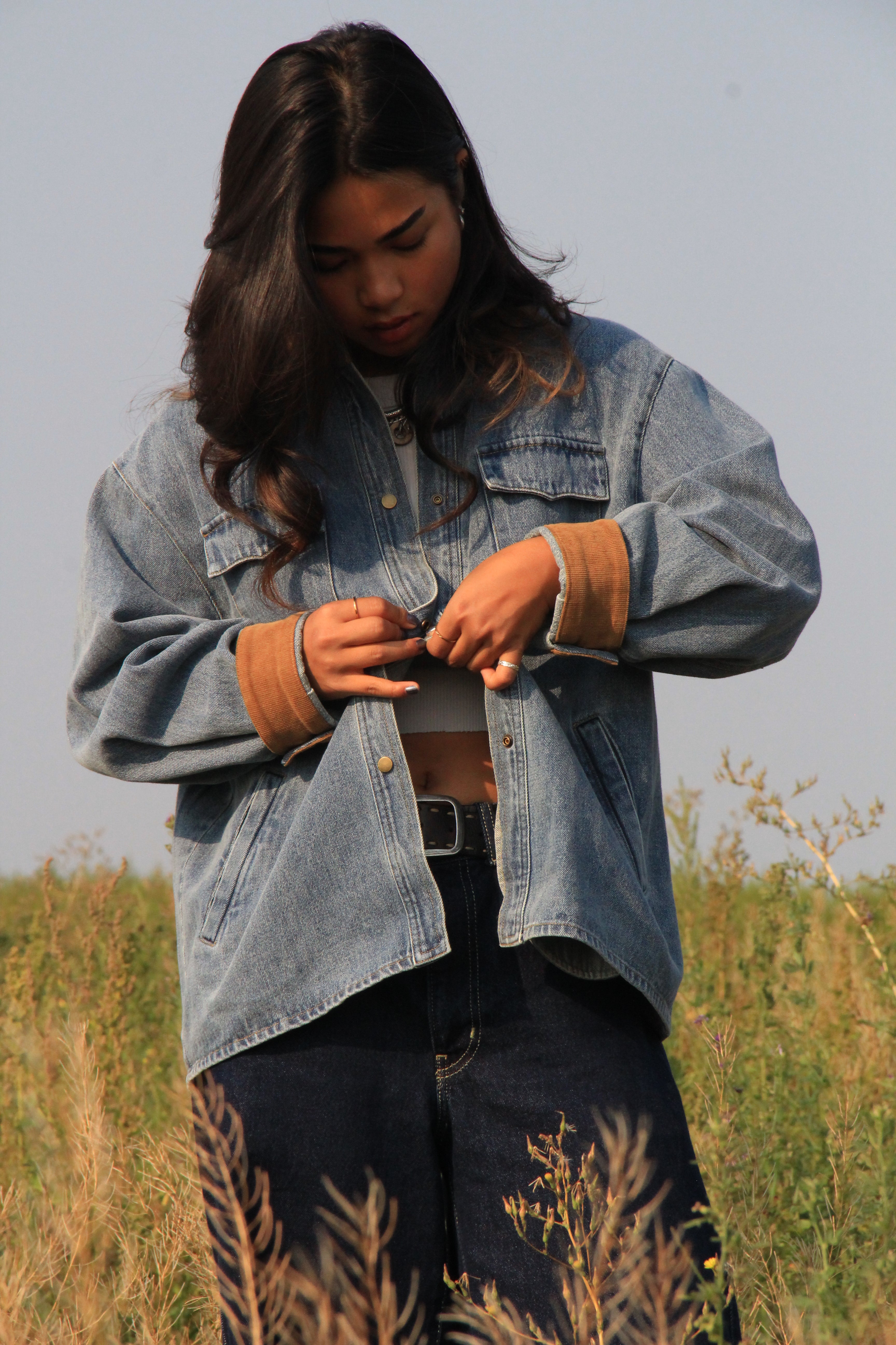A woman standing in an outdoor grassy field wearing a light-wash blue denim jacket with tan ribbed cuffs, dark jeans, and a black belt.
