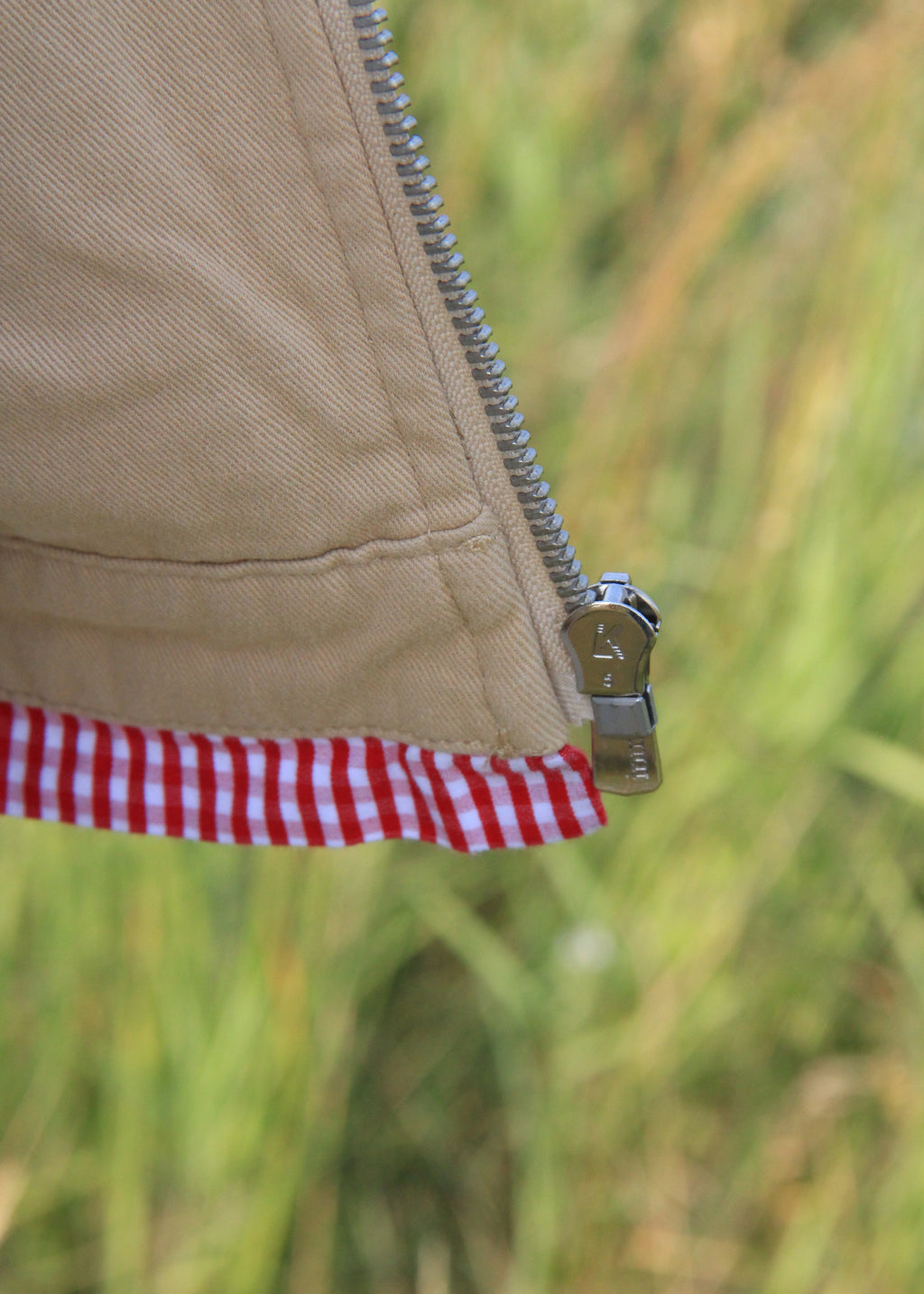 Close-up of a beige fabric zipper with a silver metal pull, showing a red and white gingham check lining along the edge, set against a blurred green and yellow outdoor background.