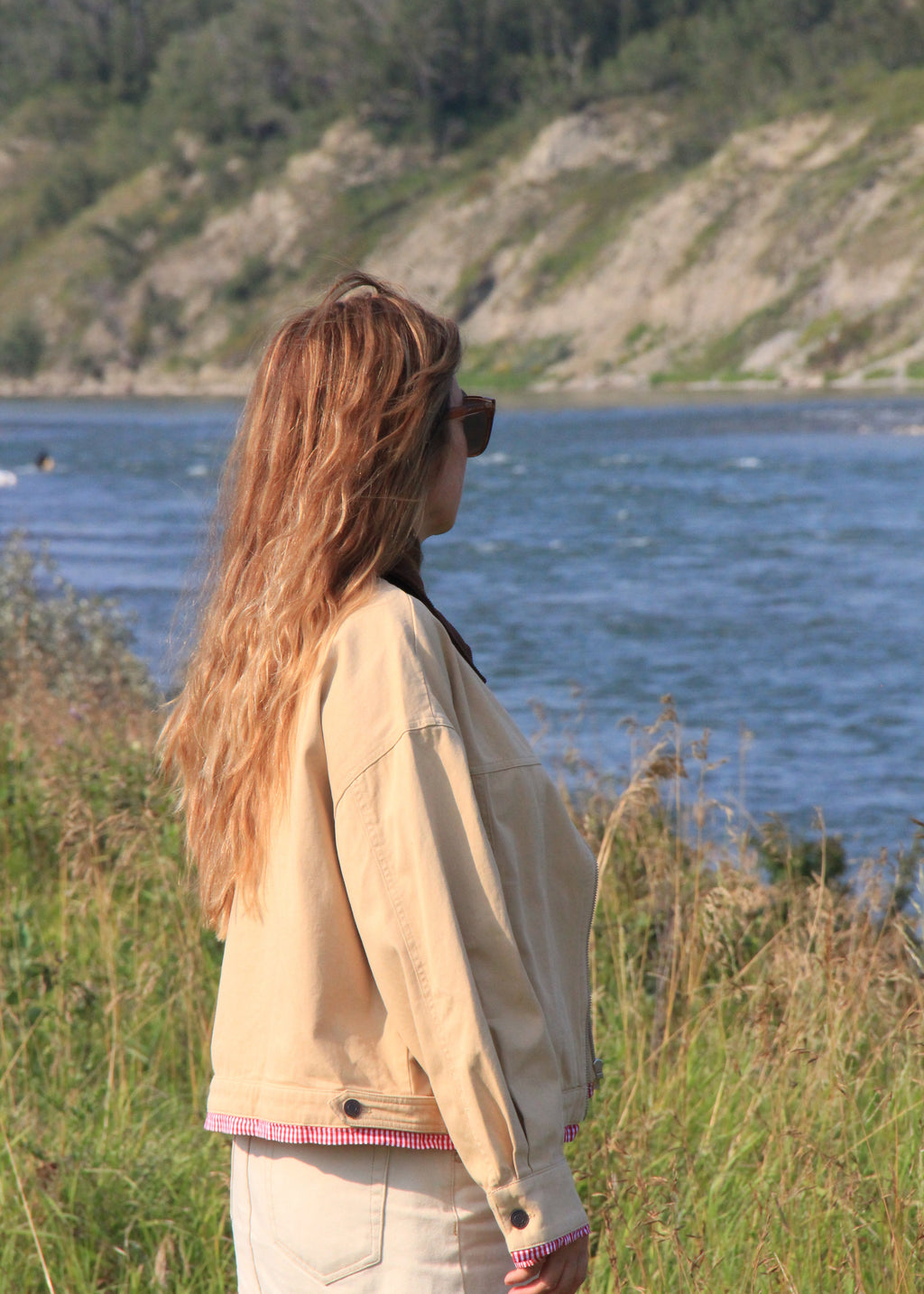 A woman with long light brown hair stands in profile outdoors by a body of water, wearing a light beige jacket with pink trim and a white skirt. Tall grasses and a rocky, tree-covered cliff are visible in the background.