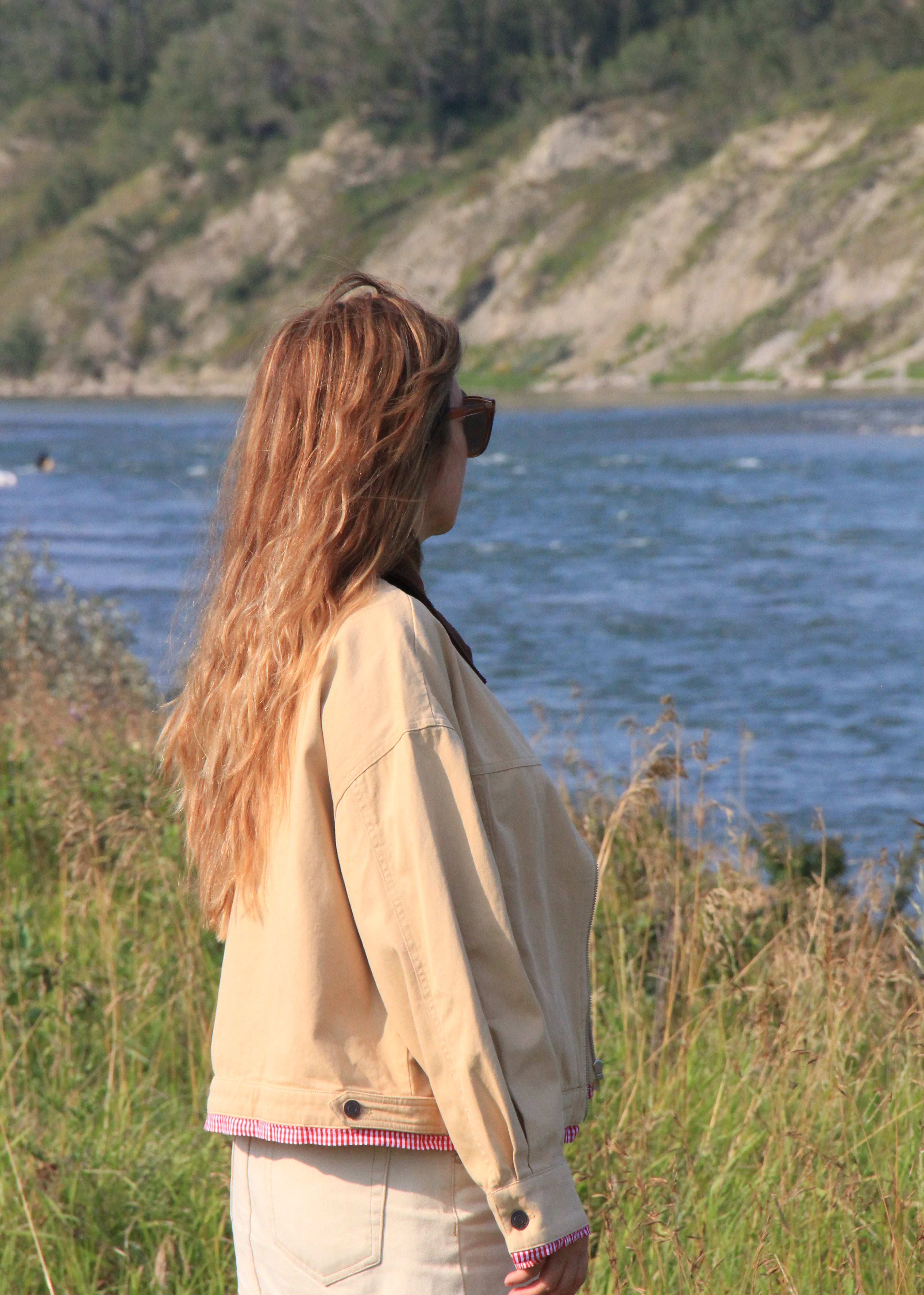 A woman with long light brown hair stands in profile outdoors by a body of water, wearing a light beige jacket with pink trim and a white skirt. Tall grasses and a rocky, tree-covered cliff are visible in the background.