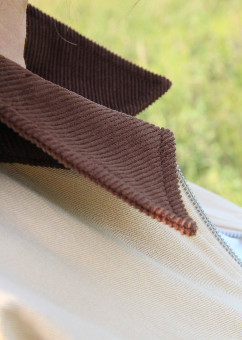 Close-up of a person wearing a light beige zip-up jacket with a wide, pointed brown corduroy collar, set against a blurred green outdoor background.