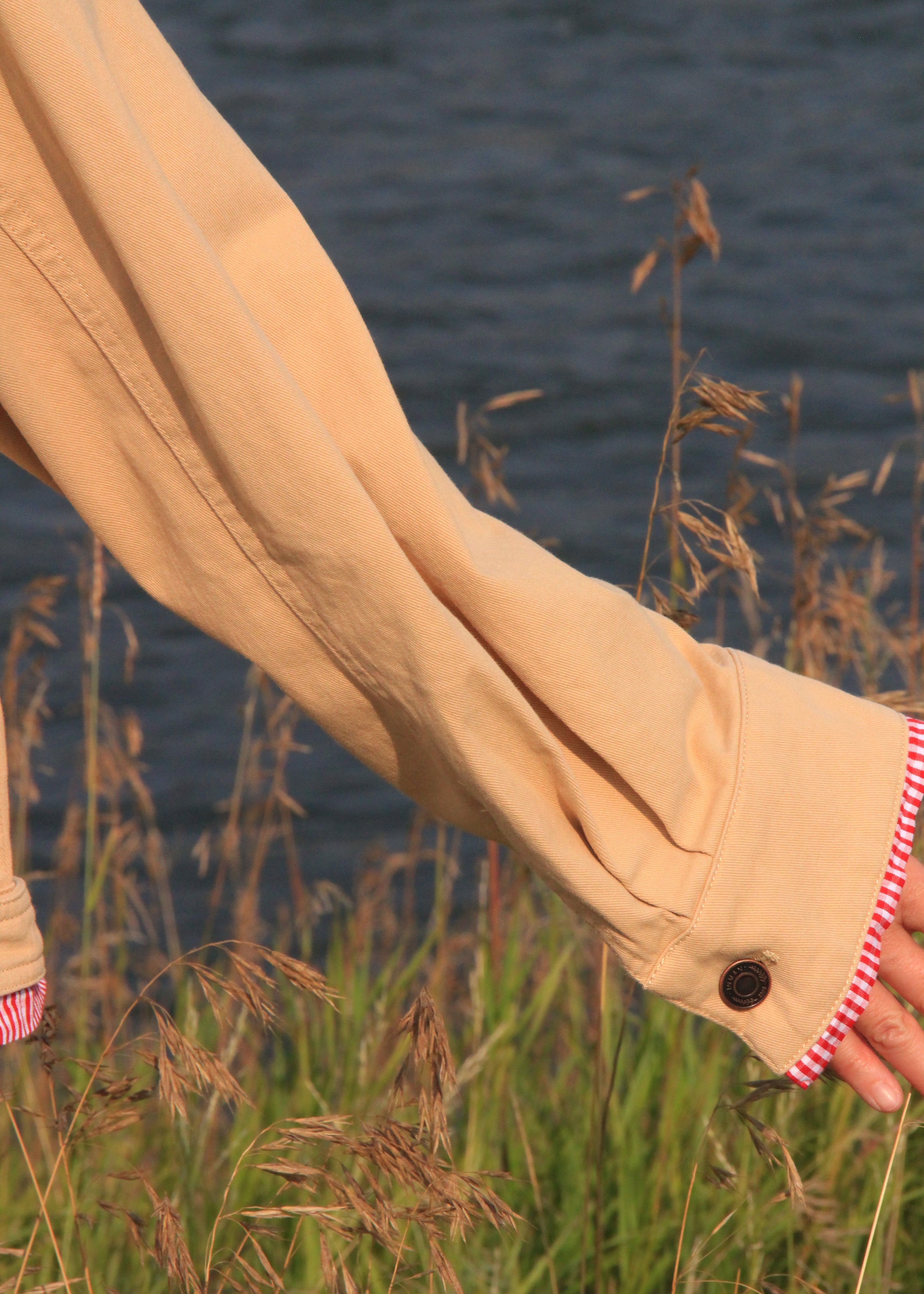 A close-up of a person's arm wearing a beige long-sleeve shirt or jacket with a buttoned cuff and a red-and-white striped fabric detail, set against a background of tall grass and water.