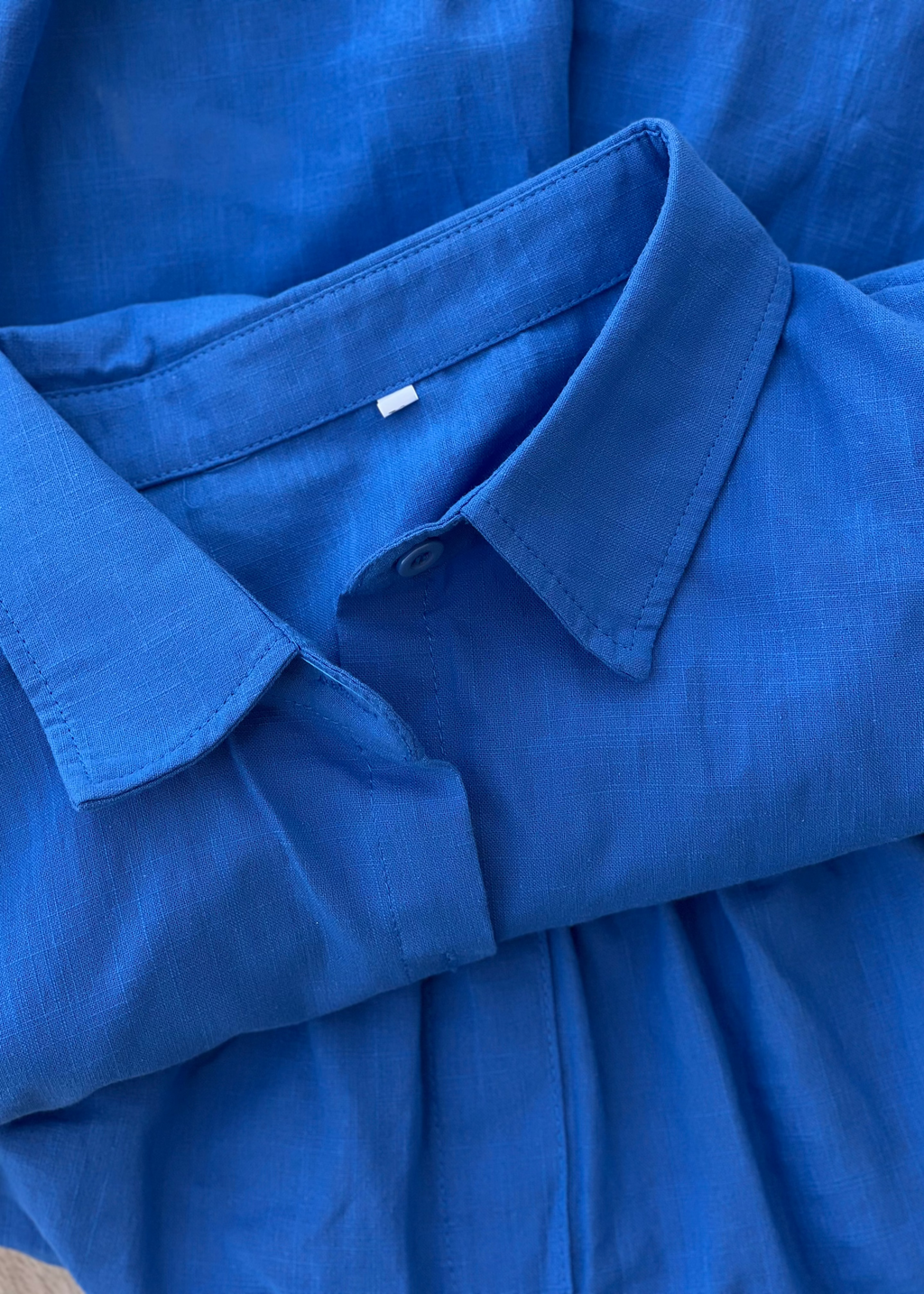 A close-up of a folded blue collared shirt or dress made from textured woven fabric, showing the collar, button placket, and soft folds.