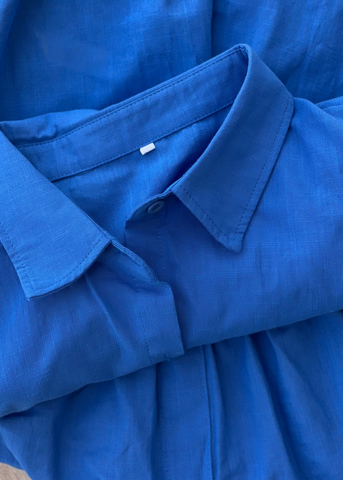 A close-up of a folded blue collared shirt or dress made from textured woven fabric, showing the collar, button placket, and soft folds.