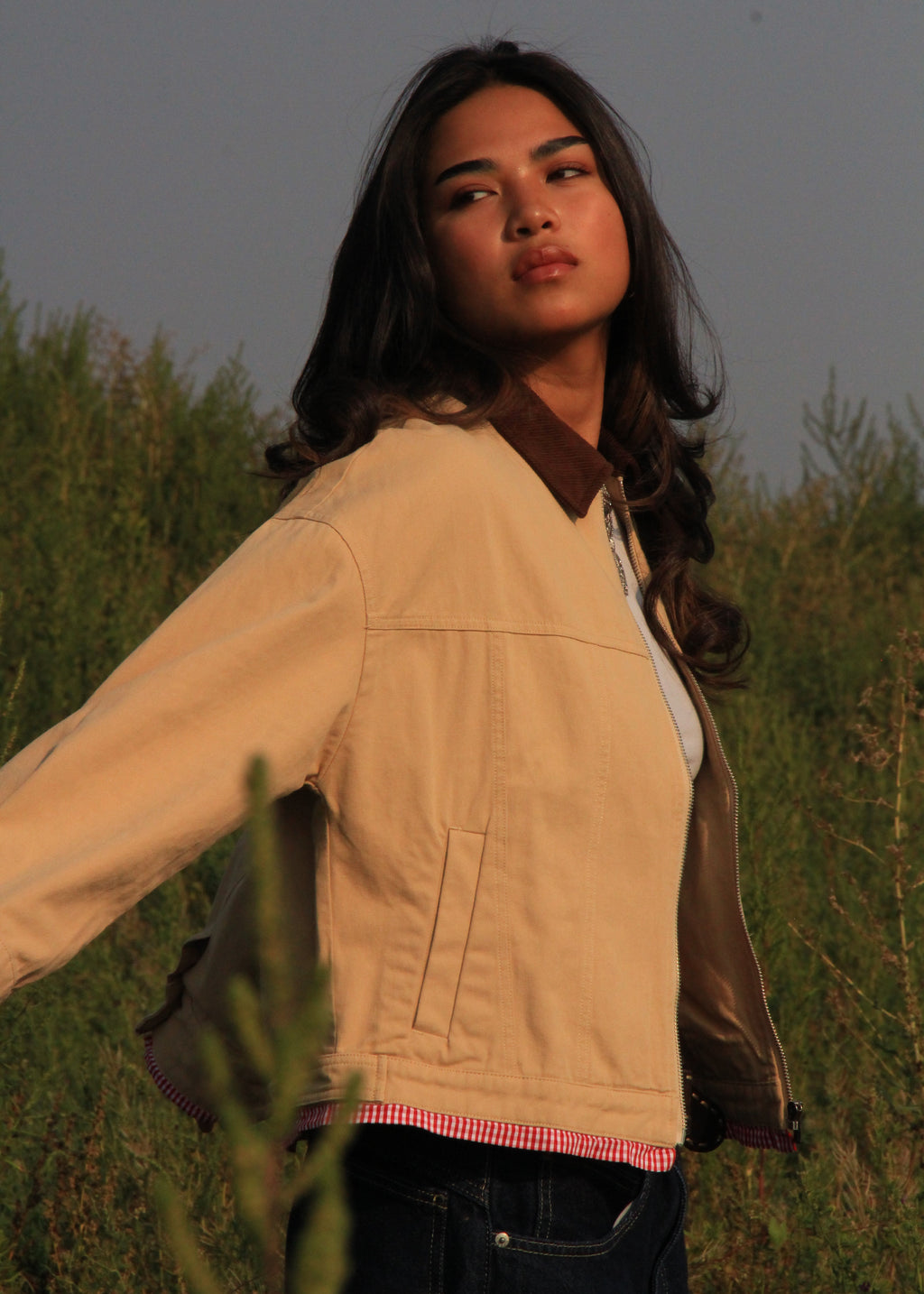A woman wearing a beige corduroy jacket with a brown collar, standing outdoors in a grassy field at sunset.