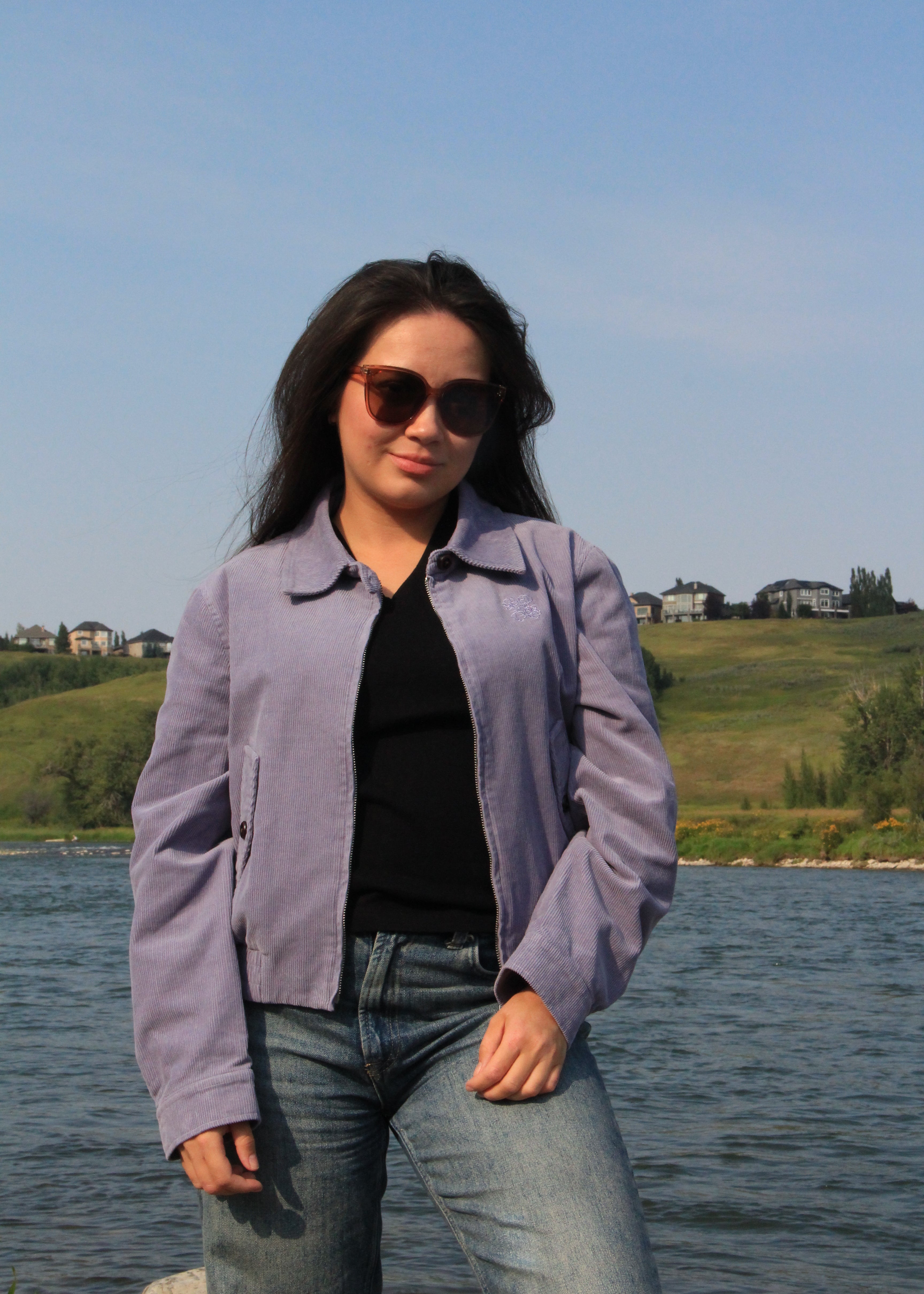 A woman wearing a light purple cropped suede jacket, black top, and blue jeans stands outdoors by a lake with hills and houses in the background.