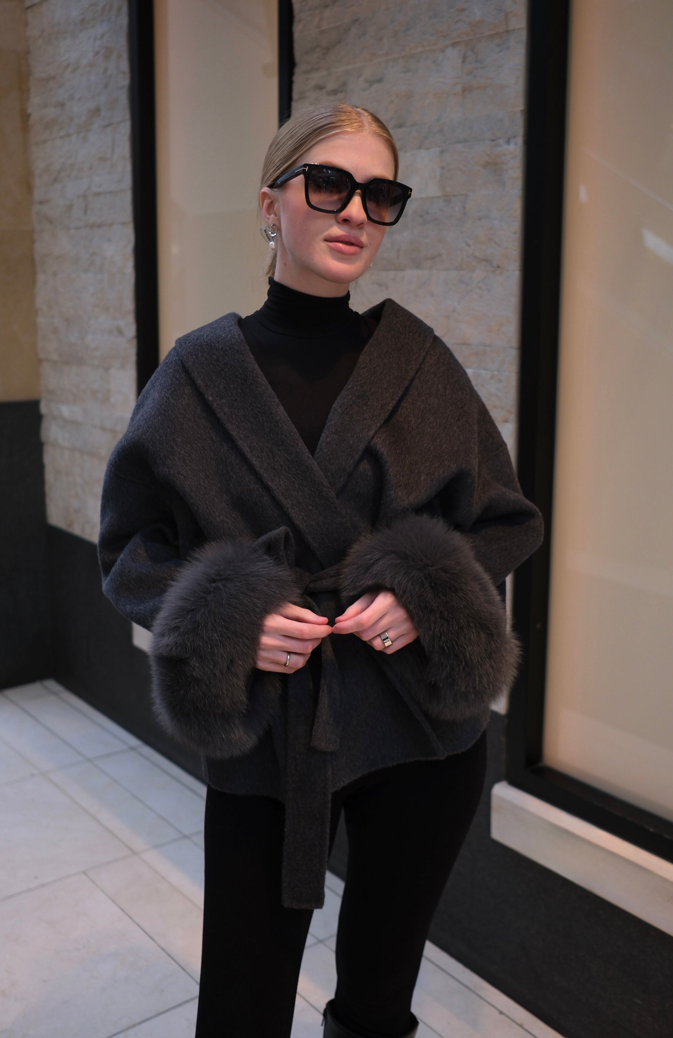 Woman wearing a charcoal grey luxury wool wrap coat with genuine rabbit fur cuffs, belted waist, oversized collar and elegant winter style.
