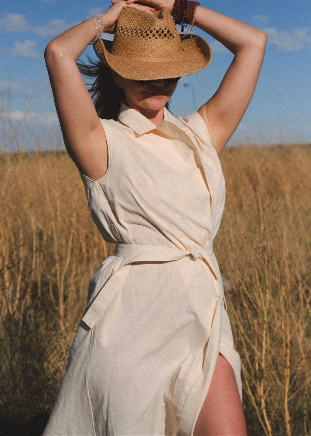 Woman in a beige dress and straw hat standing in a field with a blue sky.
