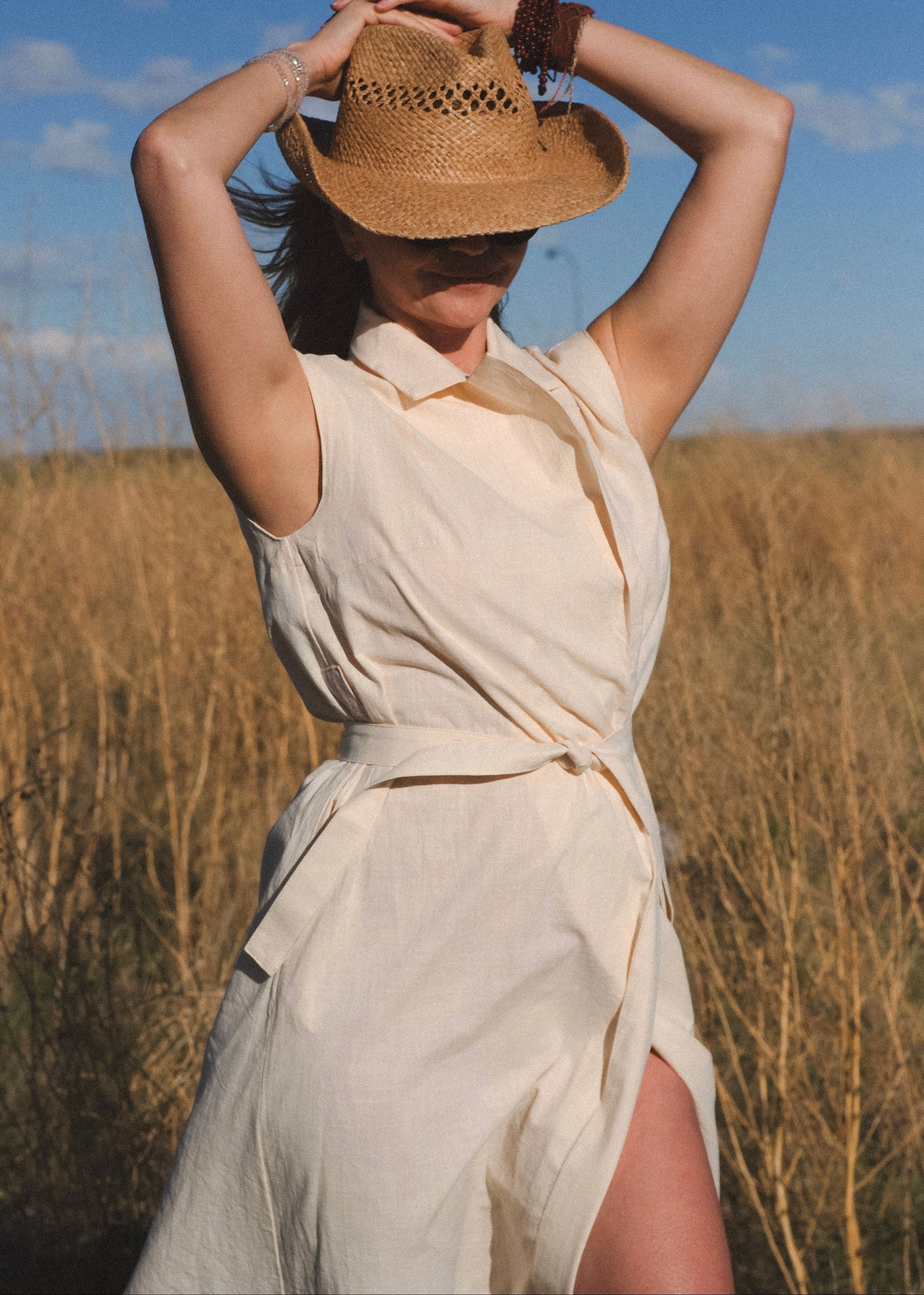 Woman in a beige dress and straw hat standing in a field with a blue sky.